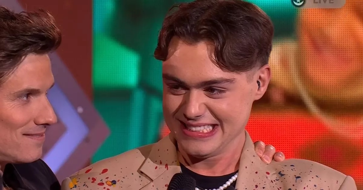 Two individuals smiling; one on the left partially visible, and the one on the right wearing a jacket with colorful splashes and a pearl necklace