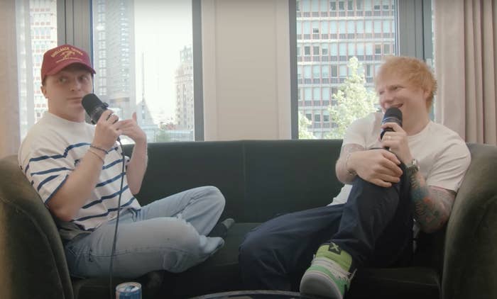 Ed Sheeran and another person sit on a couch, talking into microphones. Ed Sheeran is wearing a white t-shirt and has tattoos on his arm