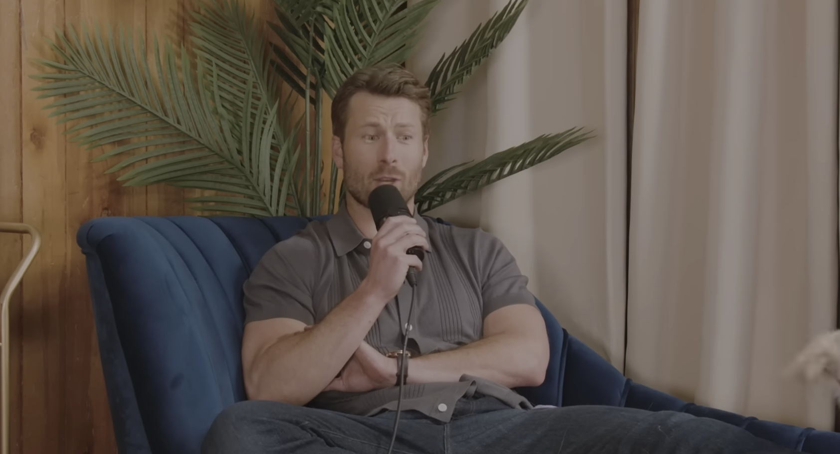 Man sitting on a blue couch holding a microphone, part of a houseplant visible behind him