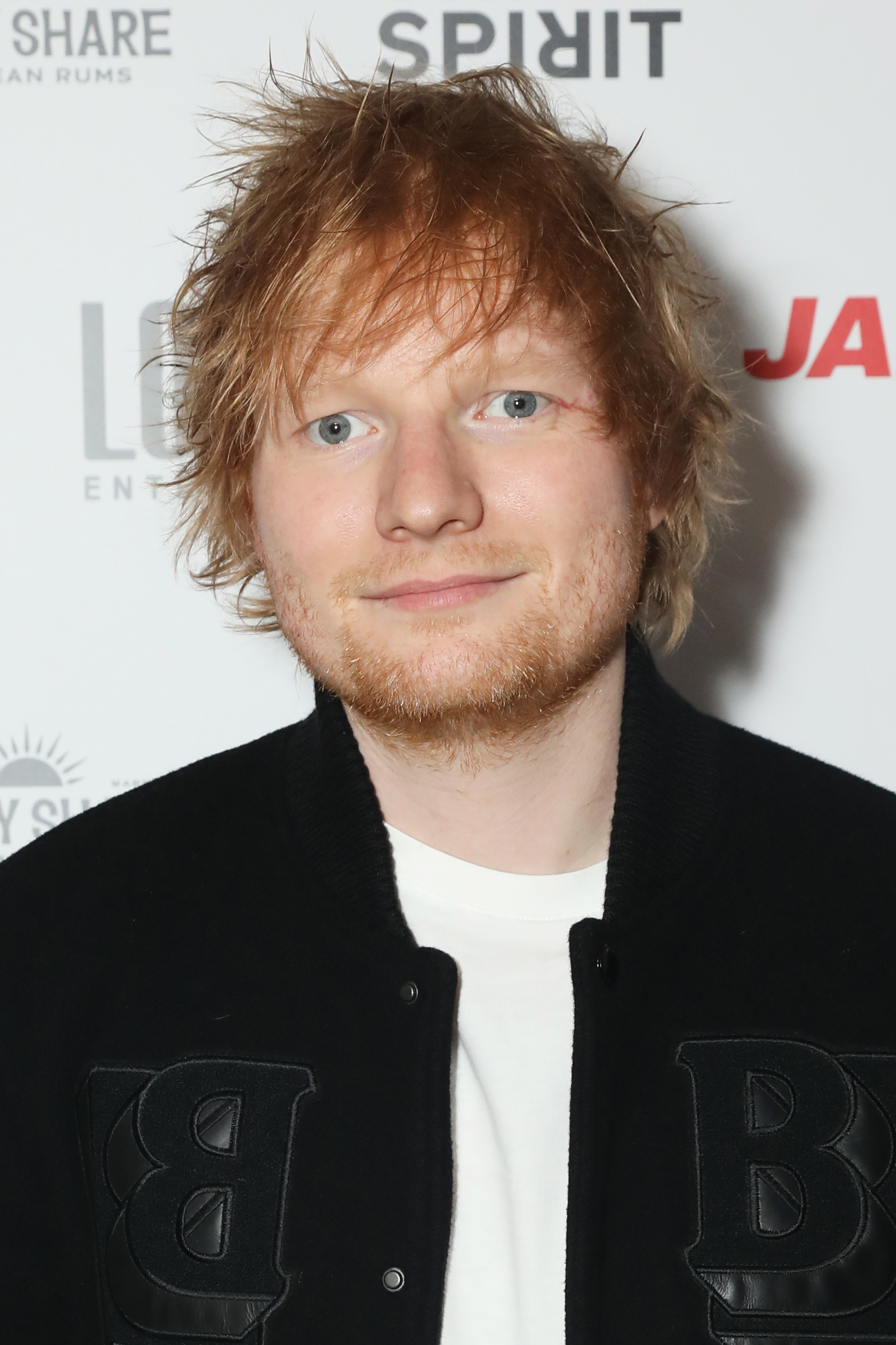 Ed Sheeran at a red carpet event wearing a casual jacket over a T-shirt, smiling at the camera