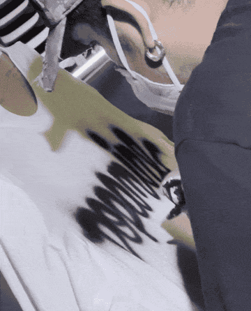 A person wearing a mask and a cap spray paints a graffiti design on a white T-shirt