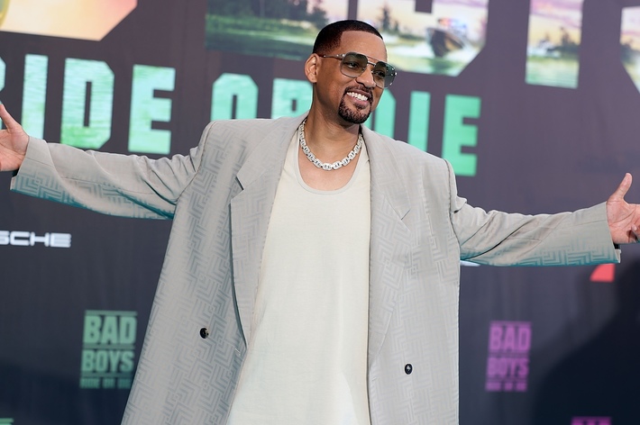 Will Smith at a movie premiere wearing a light-colored oversized suit, a white T-shirt, sunglasses, and a chain necklace, standing with arms spread open