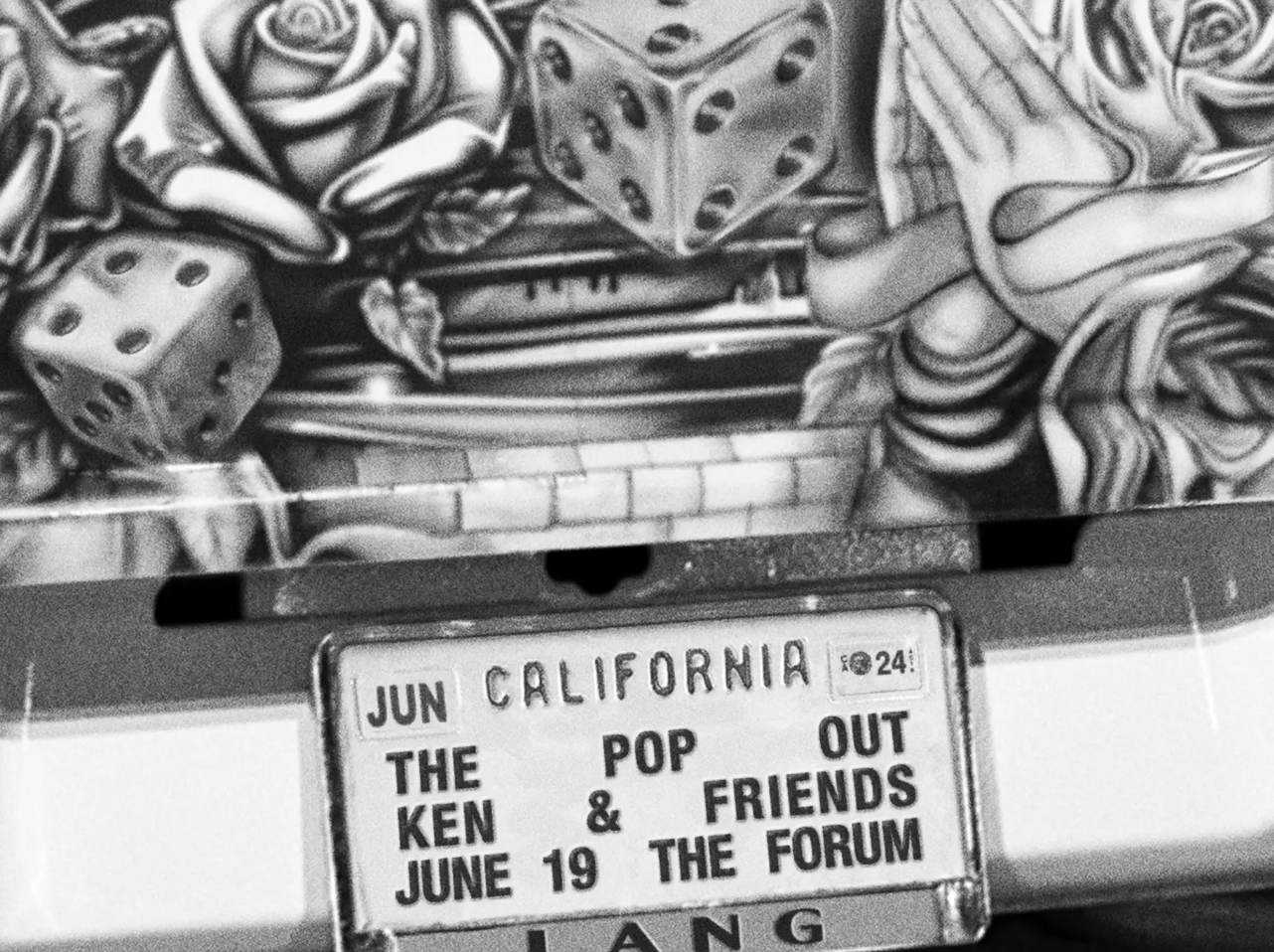 A decorative license plate on a car reads: &quot;THE POP OUT, KEN &amp; FRIENDS, JUNE 19, THE FORUM&quot;. Background has a mural with dice, roses, and praying hands