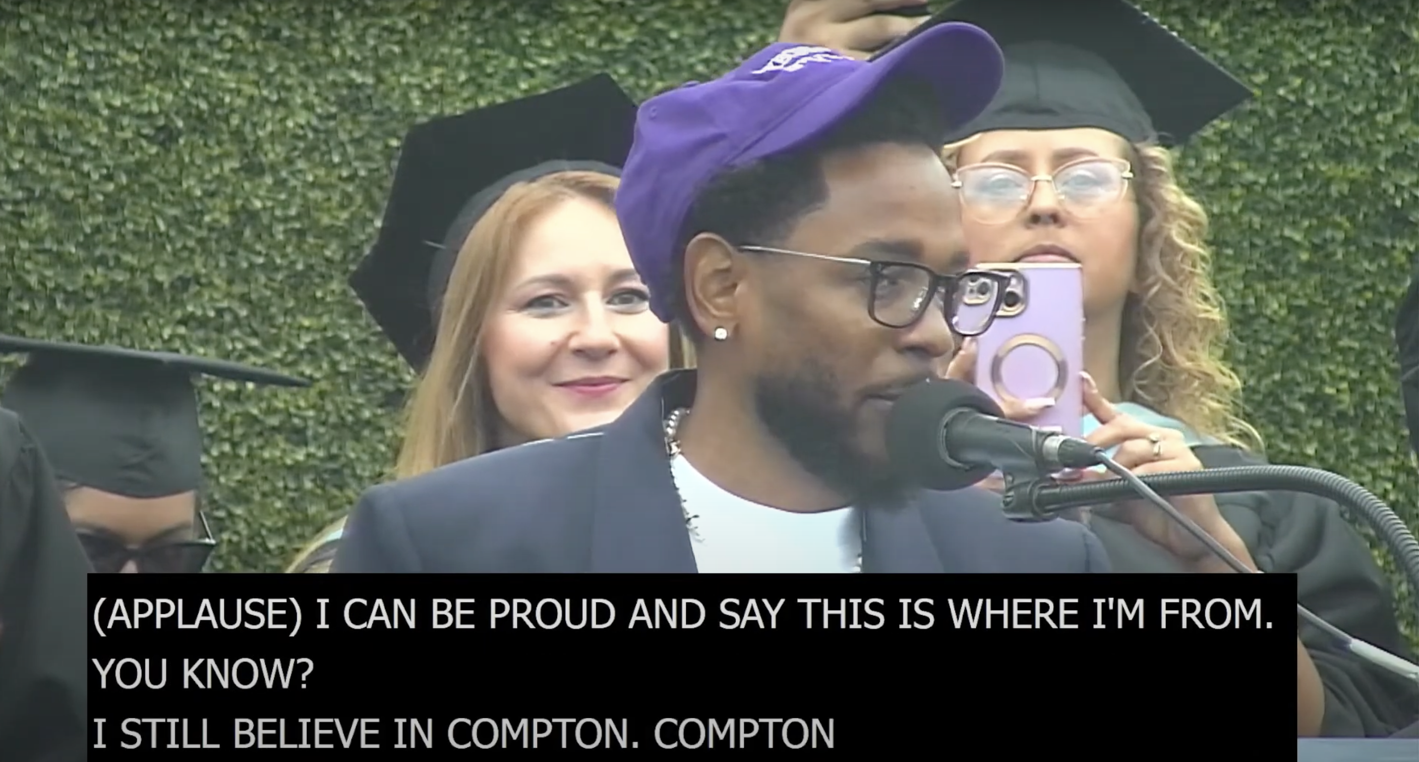 Kendrick Lamar speaks at a graduation ceremony, wearing a cap, glasses, and a purple jacket. Caption reads: &quot;(APPLAUSE) I CAN BE PROUD AND SAY THIS IS WHERE I&#x27;M FROM. YOU KNOW? I STILL BELIEVE IN COMPTON. COMPTON&quot;