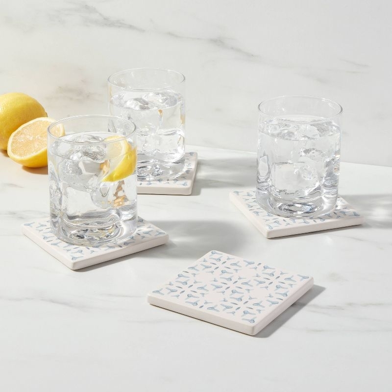 Three clear glasses with ice and a lemon slice on patterned coasters, alongside a fourth coaster and two lemons, displayed on a marble surface