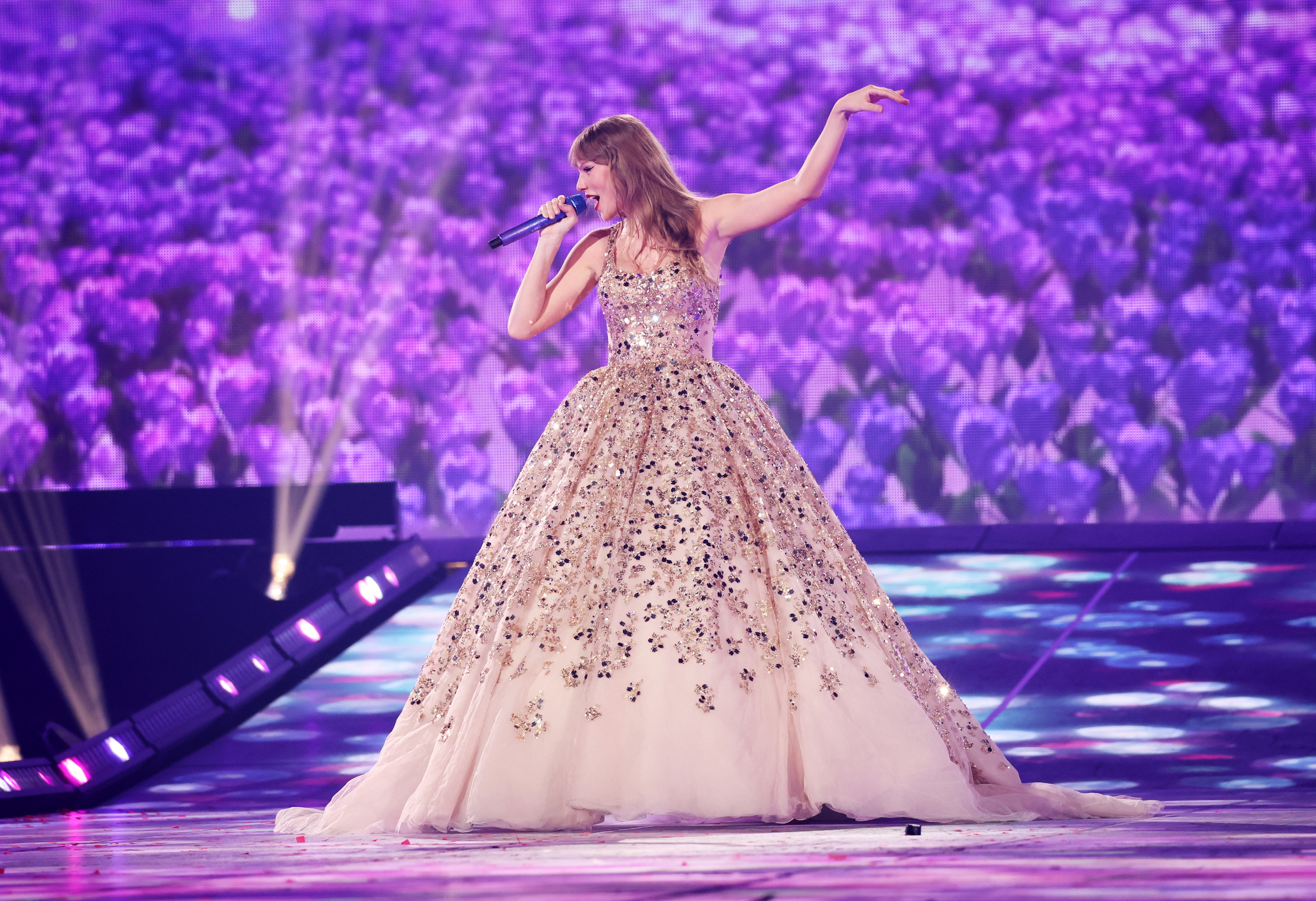 Taylor Swift is performing on stage in a sparkling, floral-patterned ball gown, singing into a microphone with a purple flower-themed backdrop behind her
