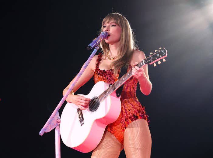 Taylor Swift performs on stage, playing an acoustic guitar. She is wearing a sparkly leotard