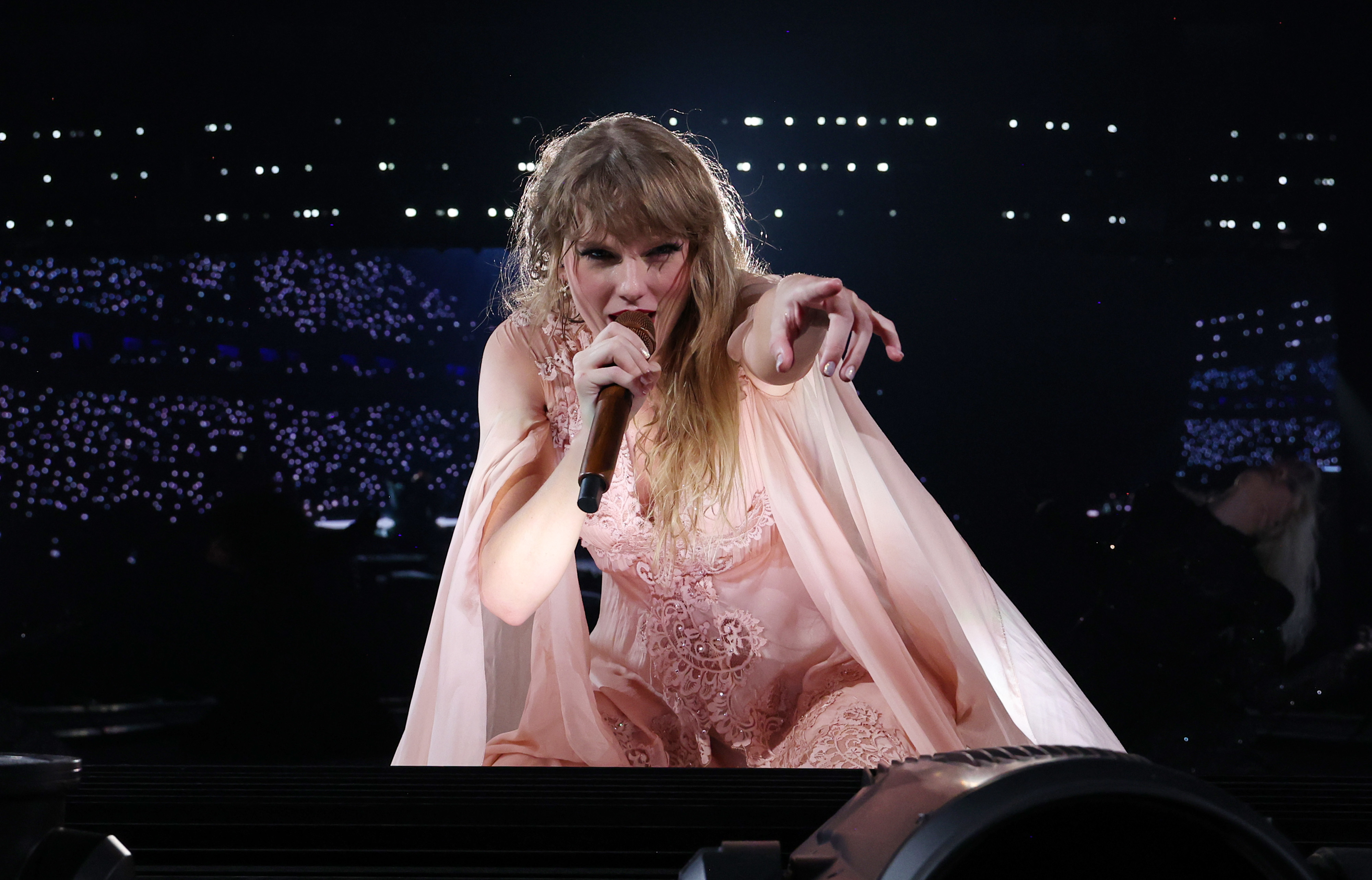 Taylor Swift performs on stage, wearing a flowing, delicate outfit, while pointing at the audience with intensity