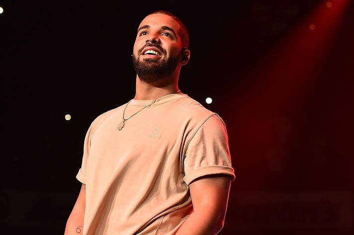 Drake smiles on stage, wearing a casual short-sleeved shirt and necklace