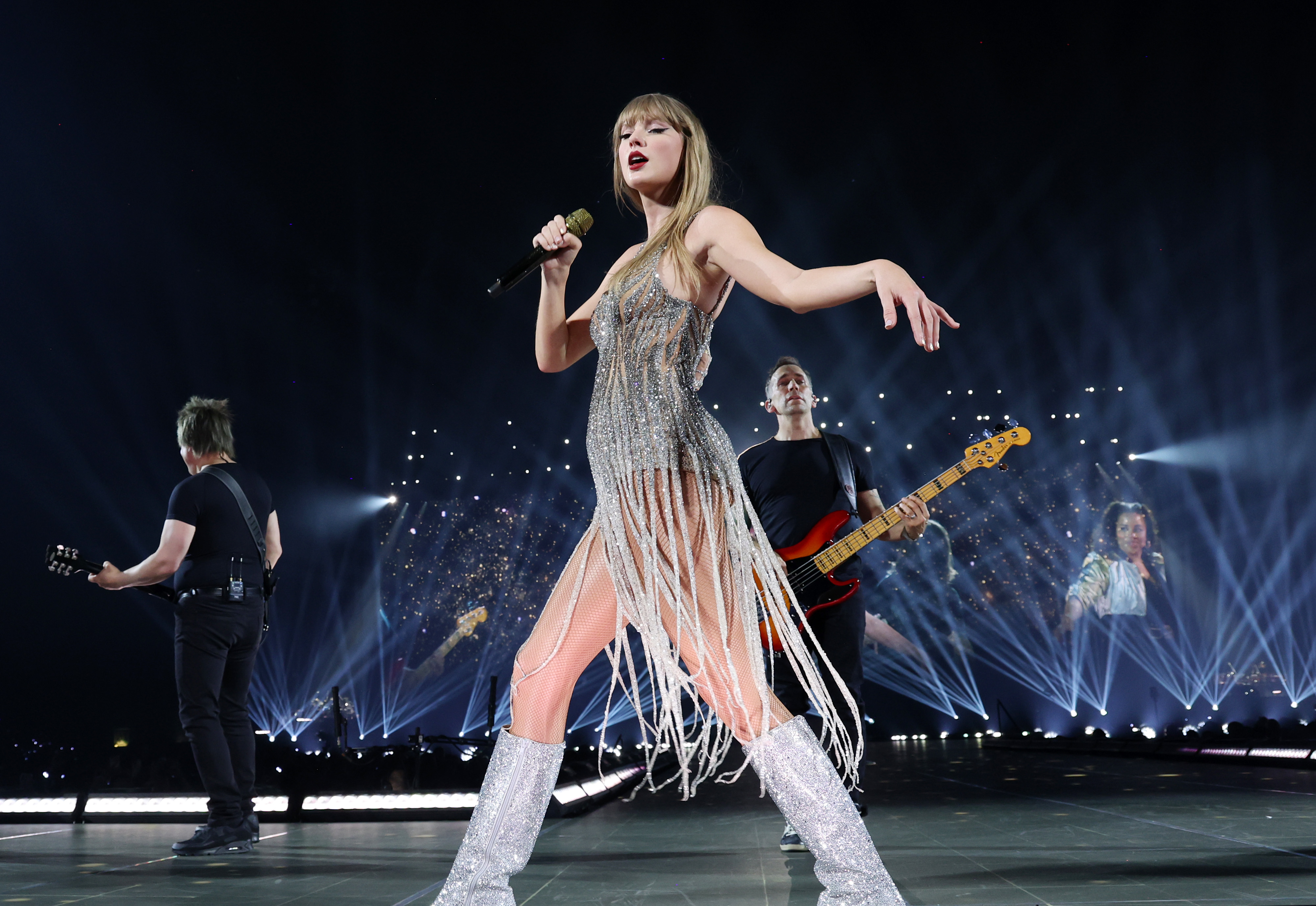 Taylor Swift performs on stage in a fringe dress, accompanied by two guitarists, during a concert. Bright stage lights and large screen projections enhance the scene