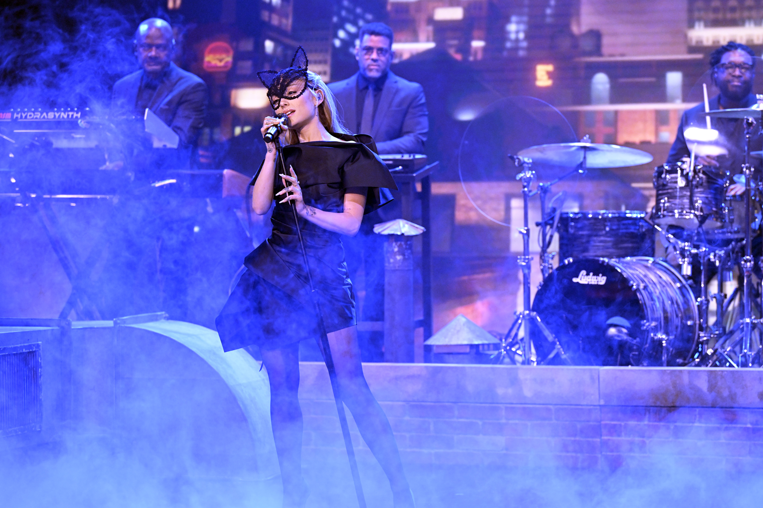 Ariana Grande performs on stage in a black dress and lace mask, accompanied by a band, with a cityscape backdrop and stage smoke effects