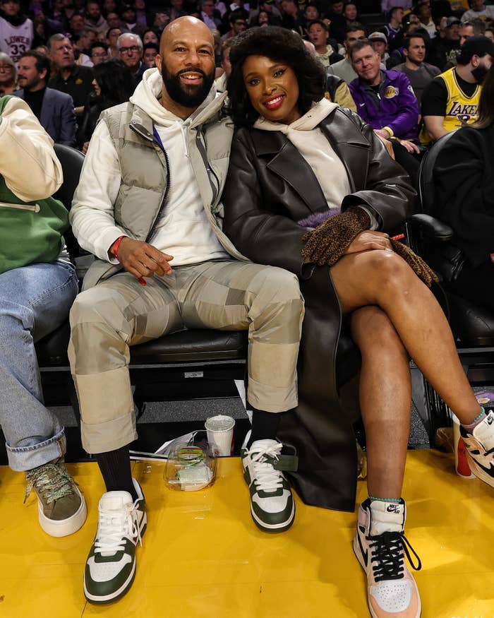 Common and Jennifer Hudson sit courtside at a basketball game. Common wears a camo outfit with a puffer vest, and Jennifer wears a long leather coat with a hoodie