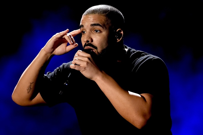 Drake performs on stage, holding a microphone close to his mouth with one hand and pointing to his head with the other