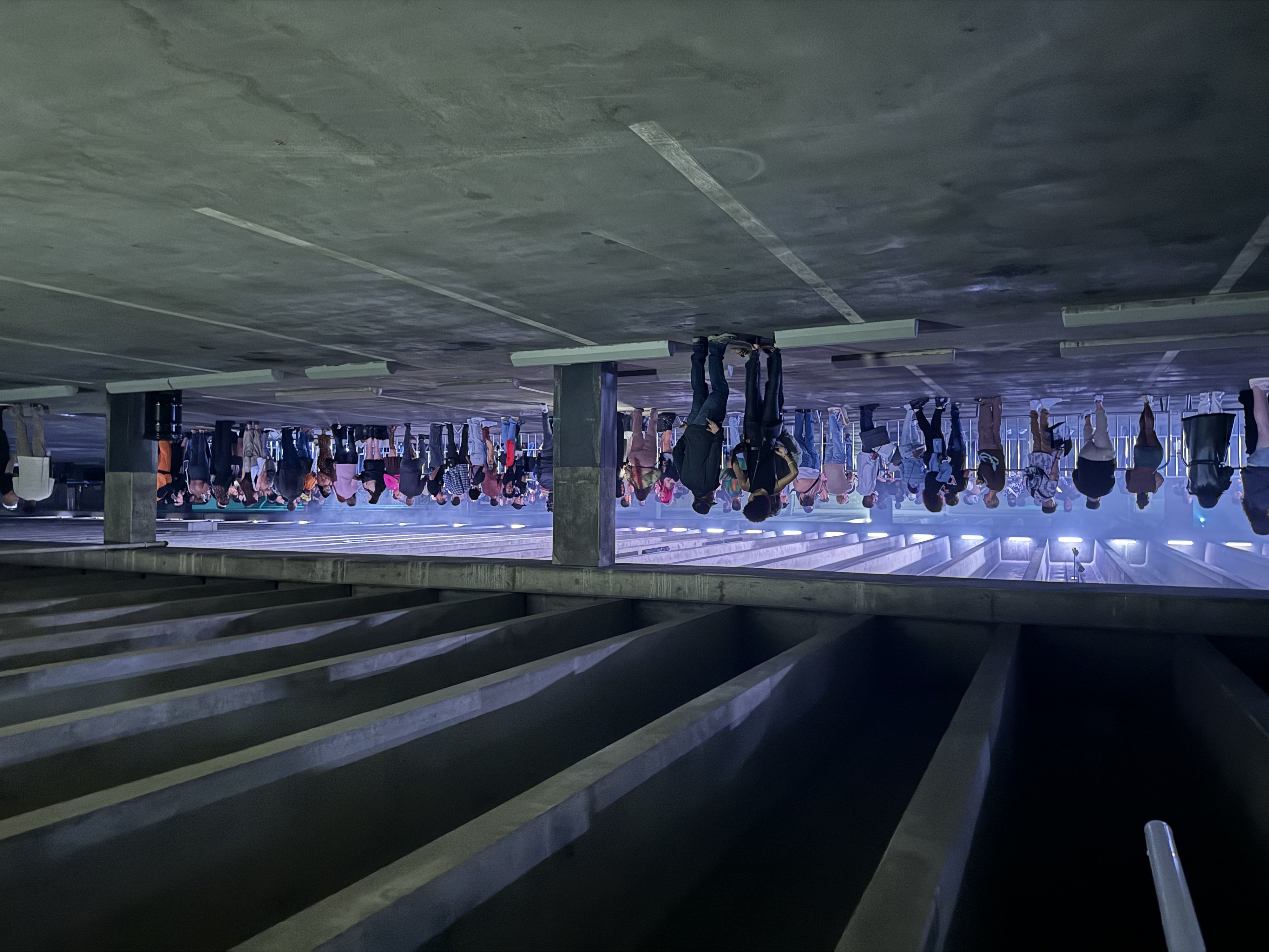 People gather in a parking garage, standing in groups, talking, and looking at their phones. The ceiling has exposed beams.