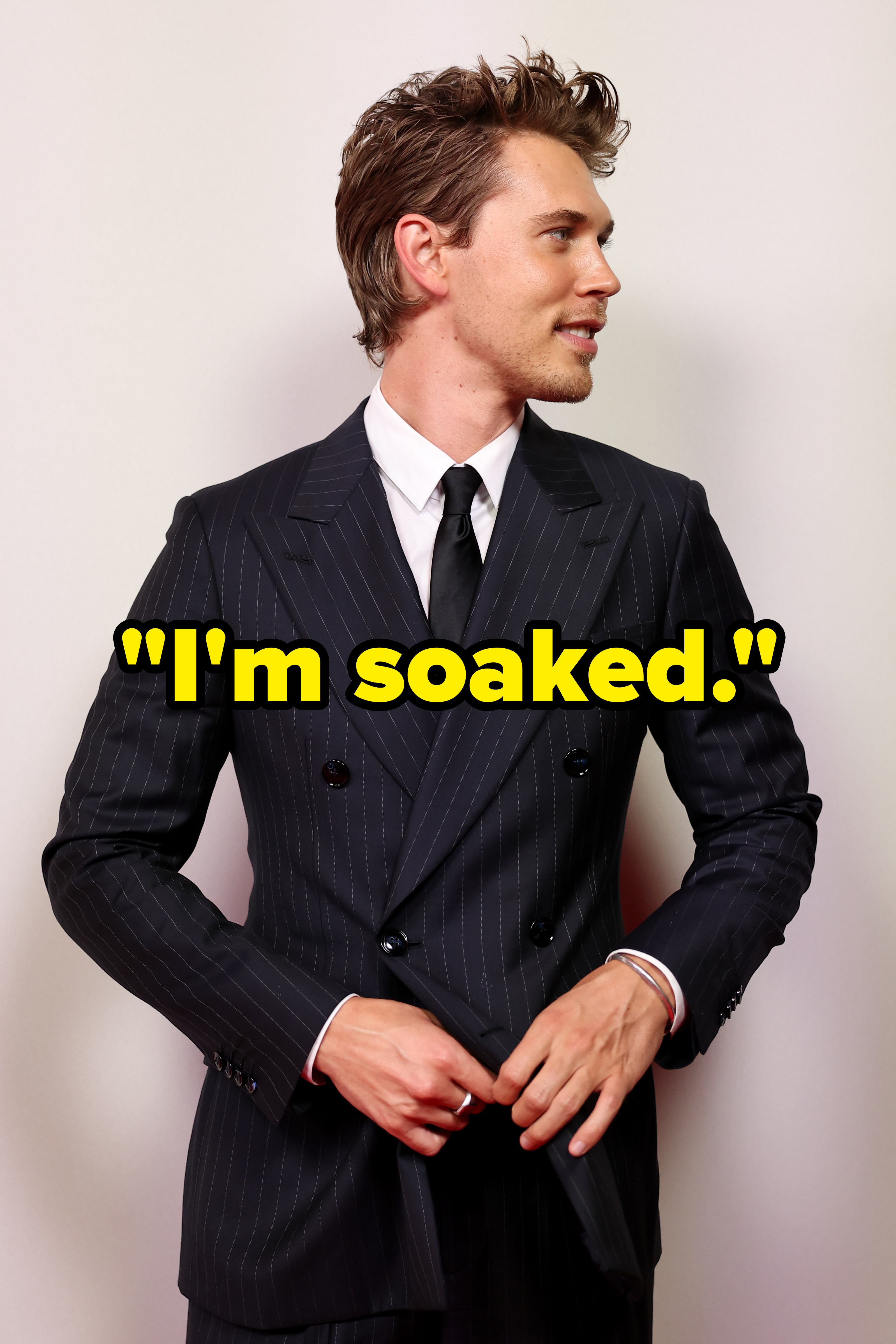 Austin Butler is wearing a pinstripe suit and a black tie, standing in front of a plain background. He is looking to the side and adjusting his suit jacket