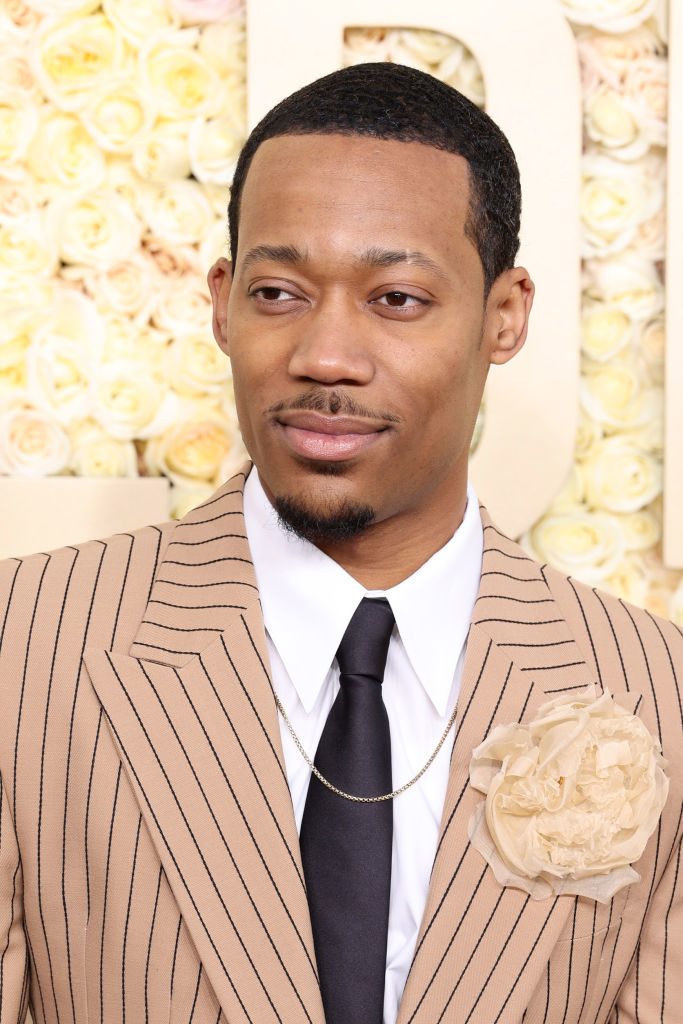 Tyler James Williams wearing a striped suit with a flower lapel pin, white shirt, black tie, and a gold chain, posing at a formal event