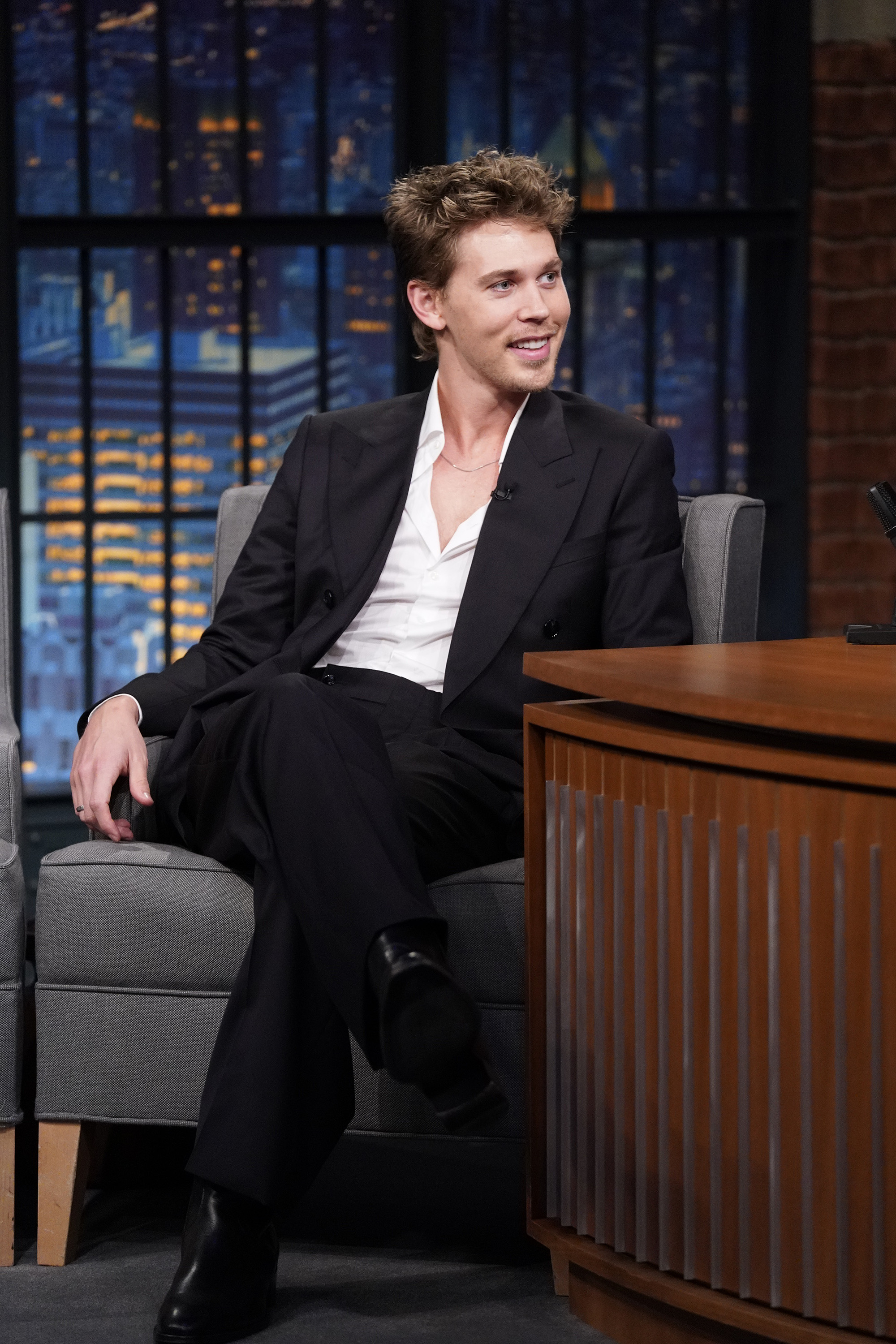 Austin Butler, in a black suit with an open-collar white shirt, sits on a talk show set, smiling and looking to his right