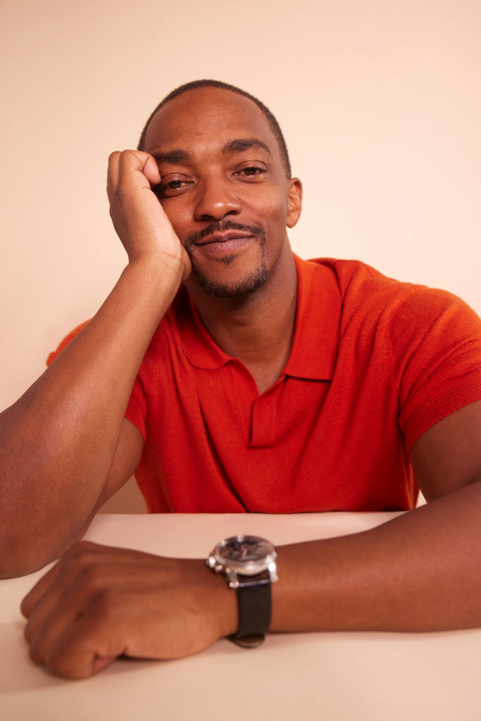 Anthony Mackie smiling and resting his head on his hand, wearing a short-sleeved, collared shirt and a wristwatch, seated at a table