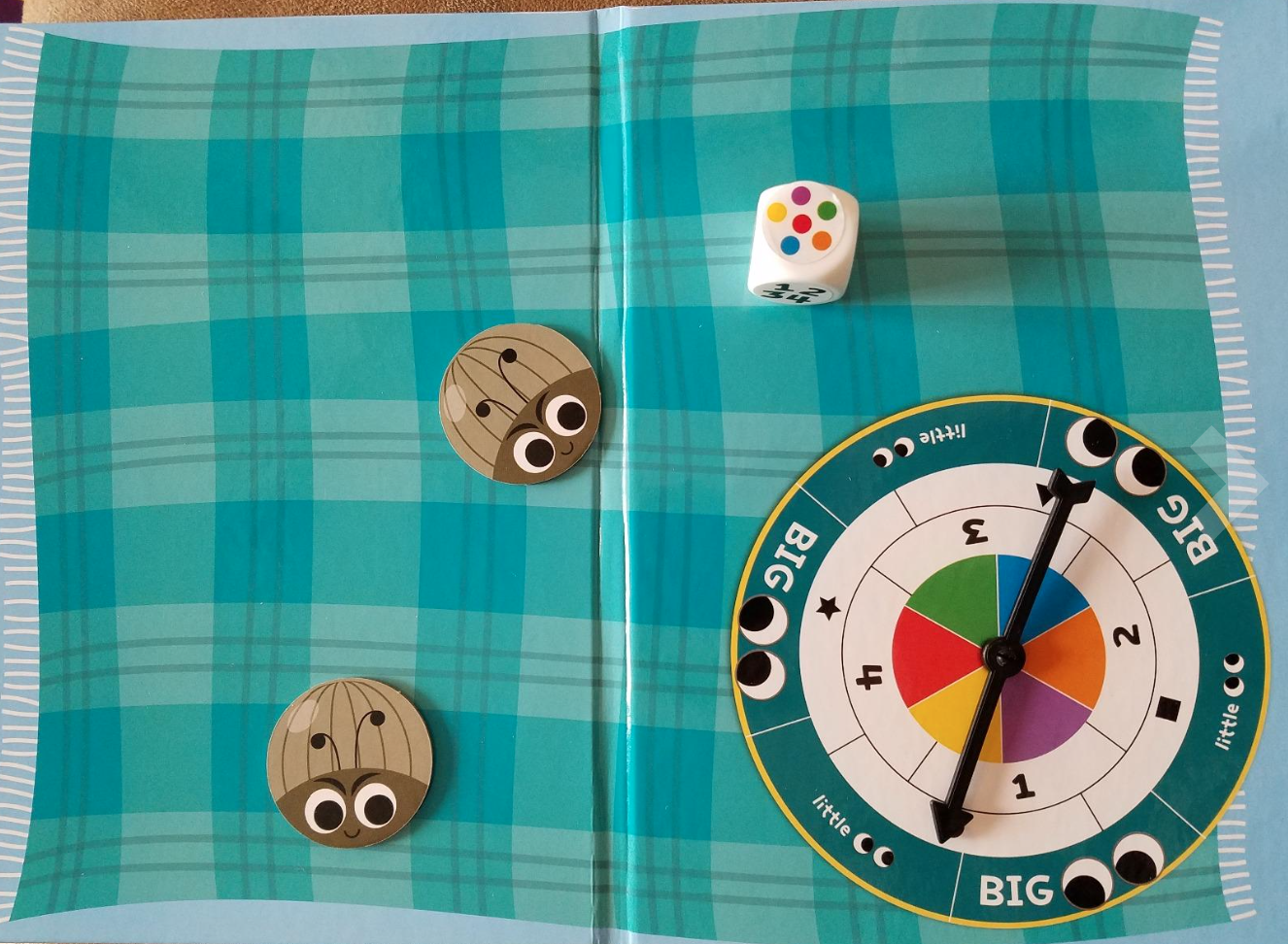 Board game setup with a colorful spinner wheel, a multi-colored die, and two tokens with cartoonish faces on a checkered background