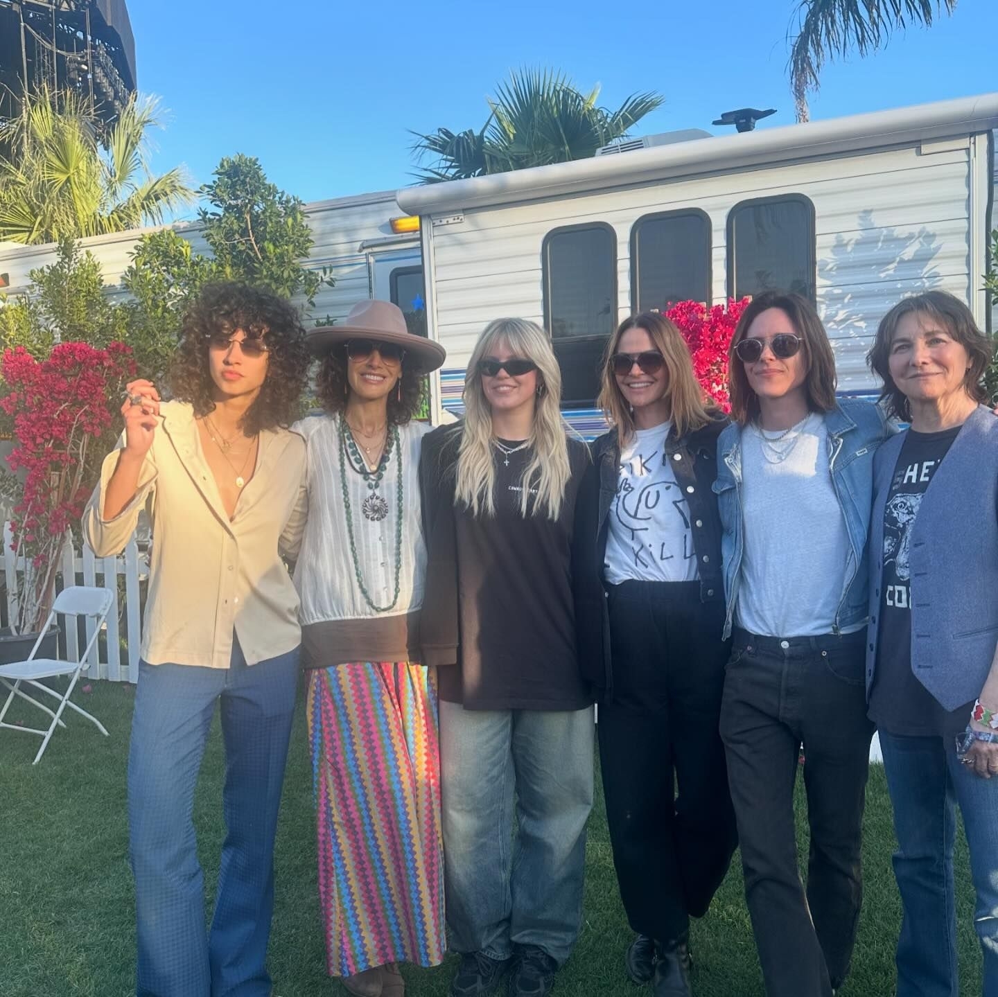 A group of six people, dressed in various styles including casual and bohemian, pose together outdoors near a trailer. Their identities are not known