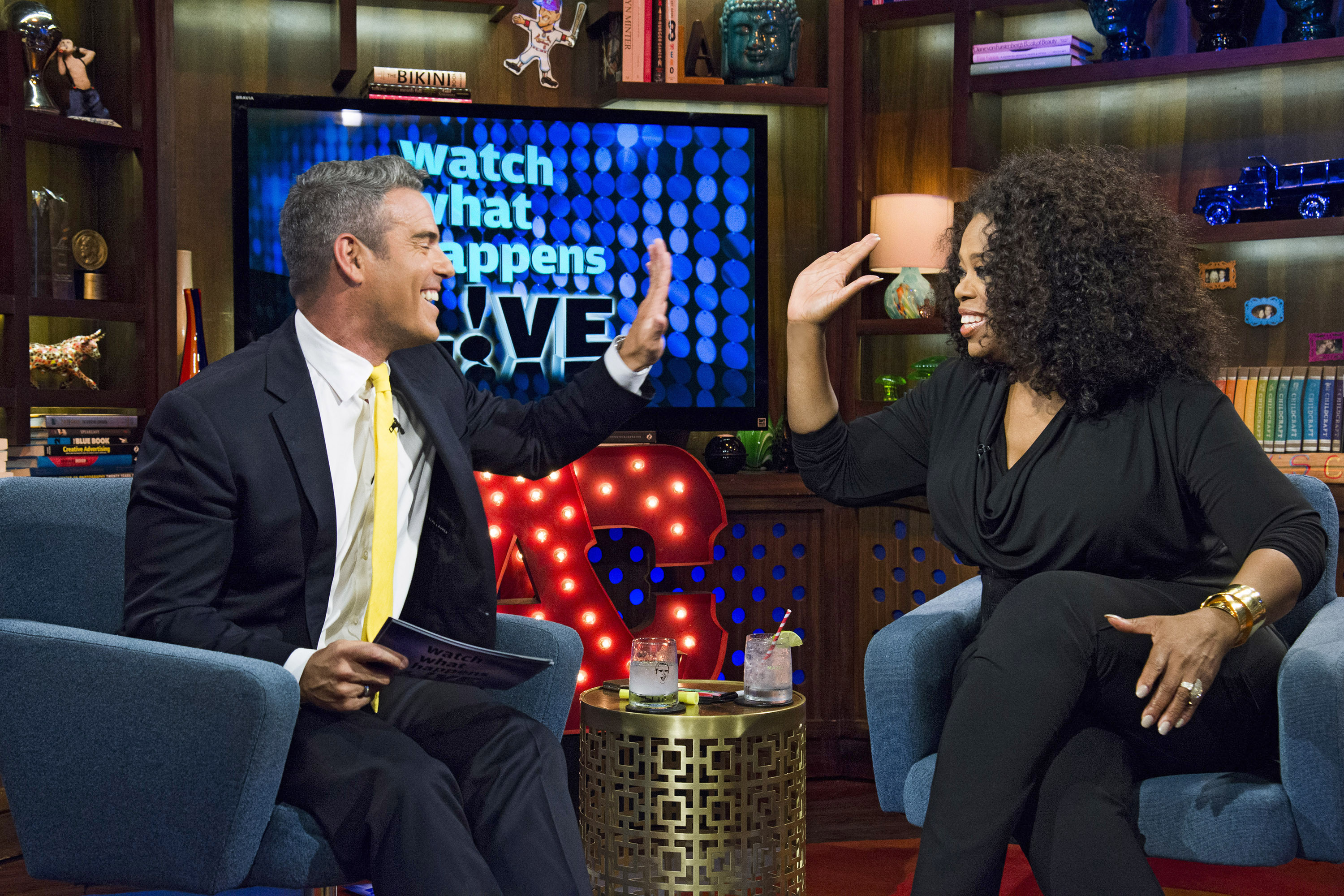 Andy Cohen and Oprah Winfrey high-fiving each other on a talk show set with bookshelves and colorful decorations in the background