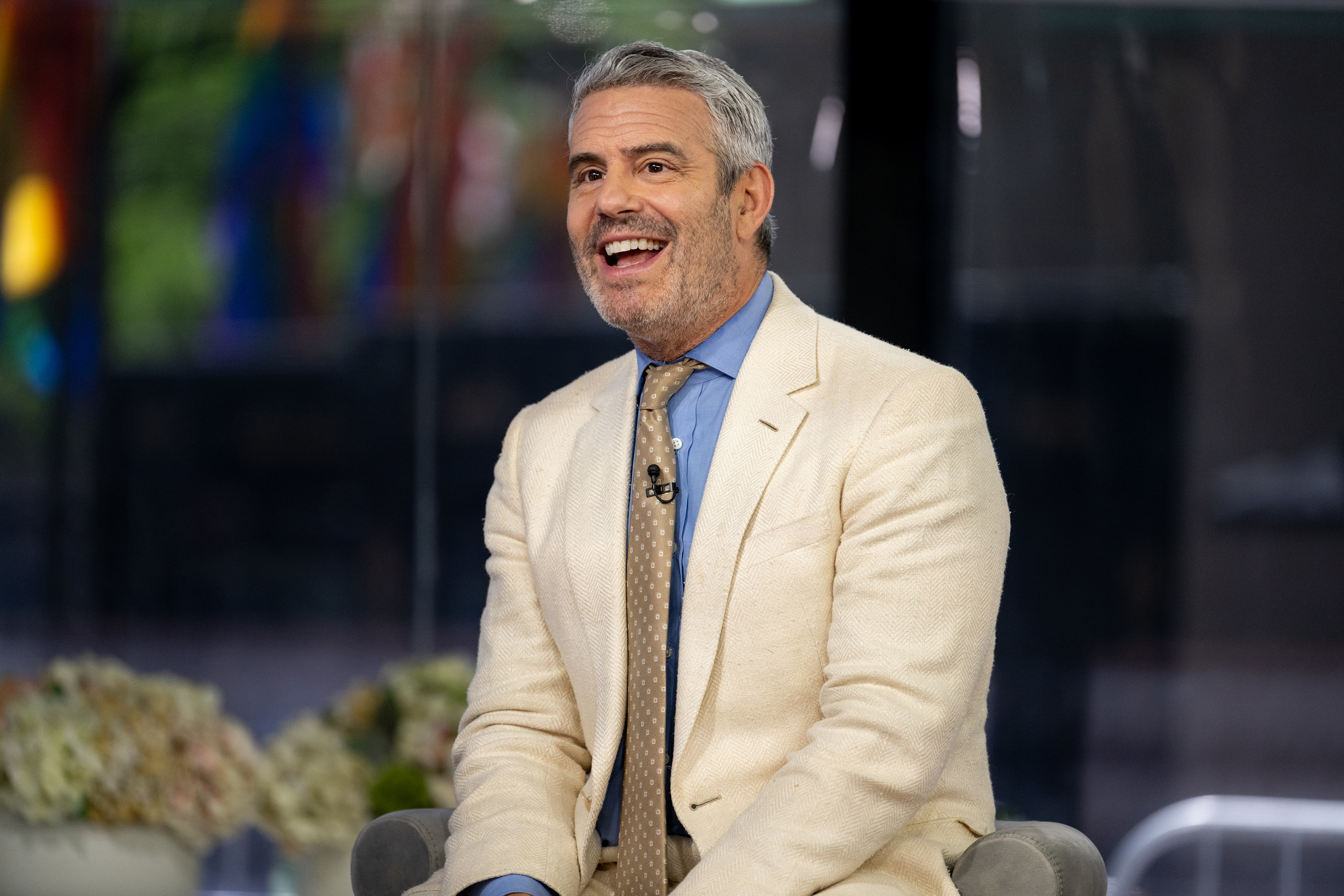 Andy Cohen is smiling, seated in an indoor setting, wearing a light-colored suit jacket, a blue dress shirt, and a patterned tie
