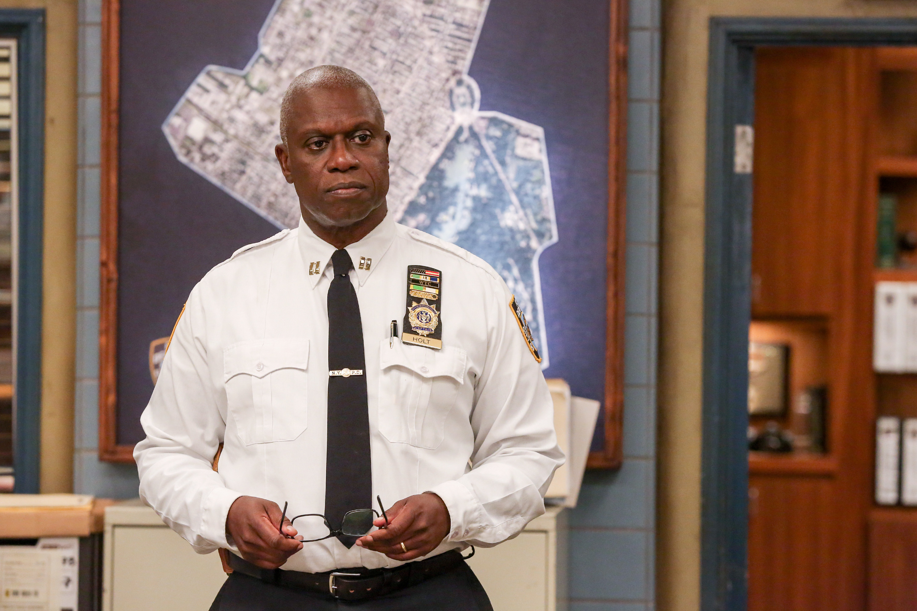 Raymond Holt in police uniform stands holding glasses in a precinct office, with a large map on the wall behind him