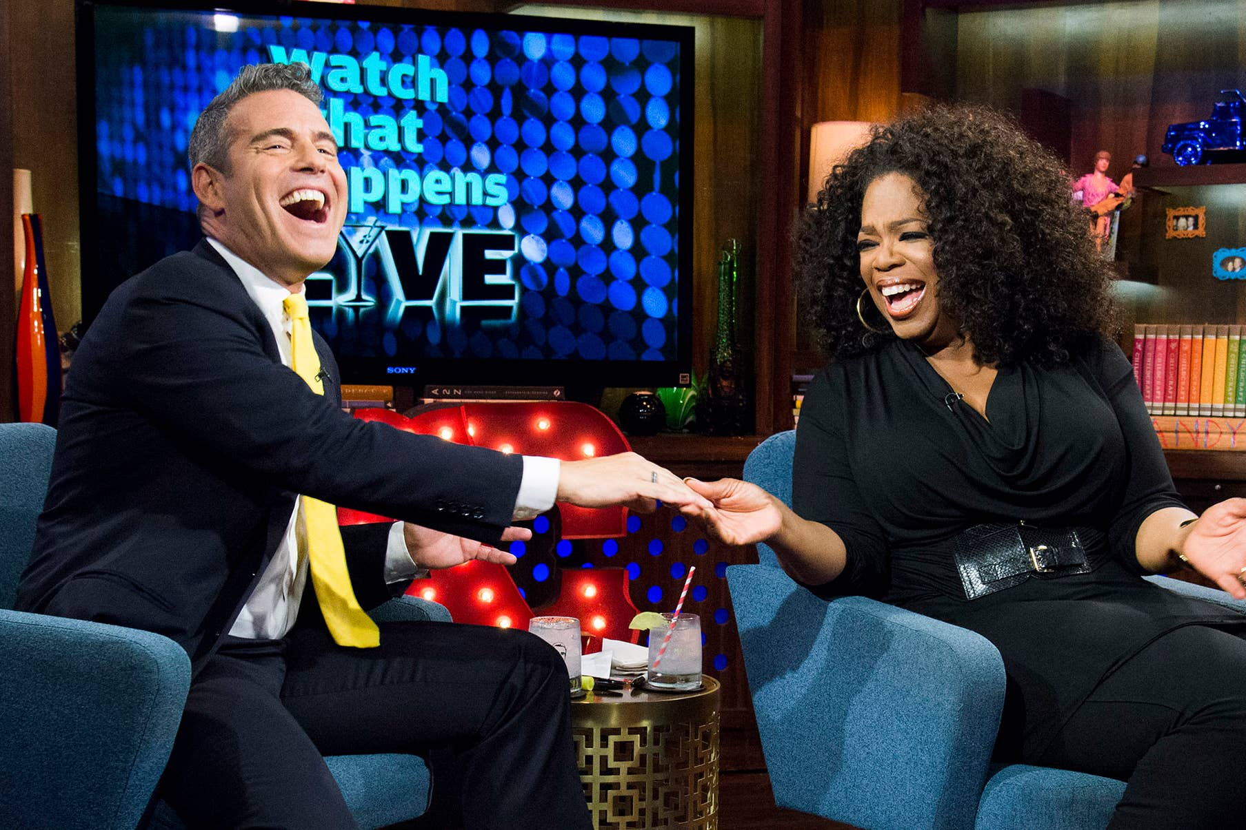Andy Cohen and Oprah Winfrey chat and laugh on a brightly decorated talk show set on "Watch What Happens Live". Andy is in a suit, Oprah wears a stylish black outfit