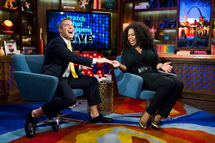 Andy Cohen and Oprah Winfrey chat and laugh on a brightly decorated talk show set on "Watch What Happens Live". Andy is in a suit, Oprah wears a stylish black outfit