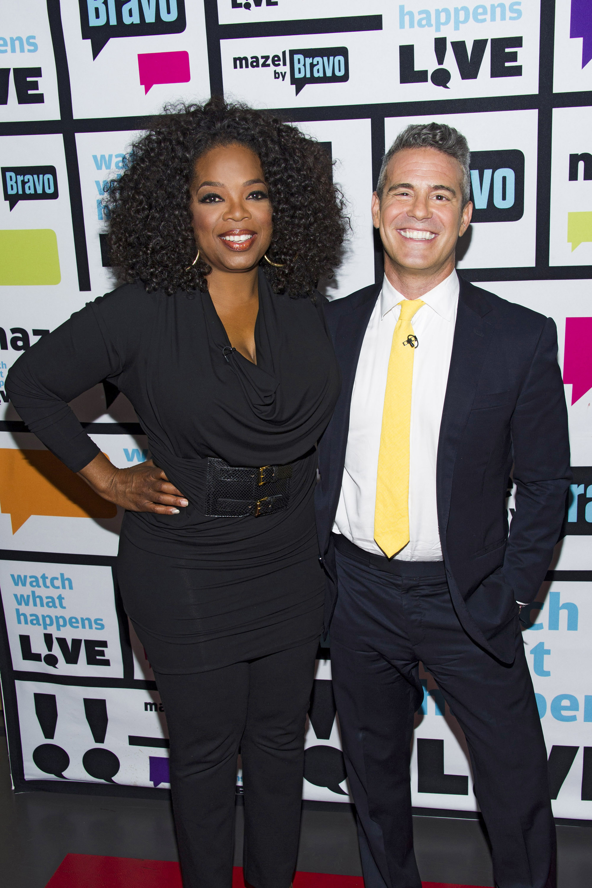 Oprah Winfrey in a stylish black outfit and Andy Cohen in a dark suit with a yellow tie smiling on the set of "Watch What Happens Live."