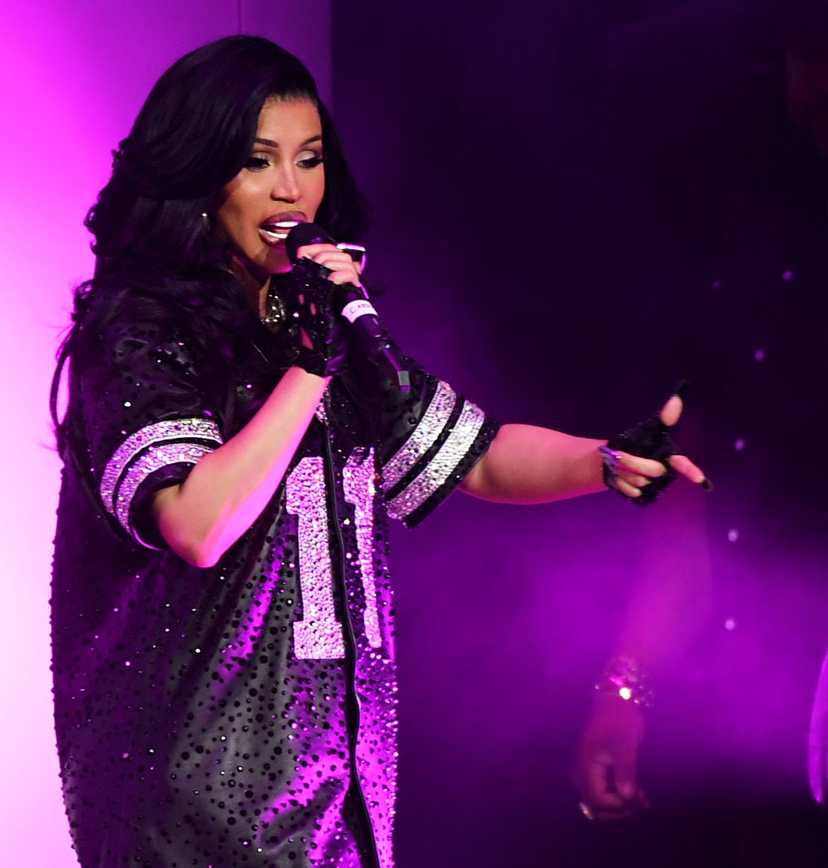 Cardi B performs on stage wearing a sequined sports jersey with the number 11. She holds a microphone and gestures while singing