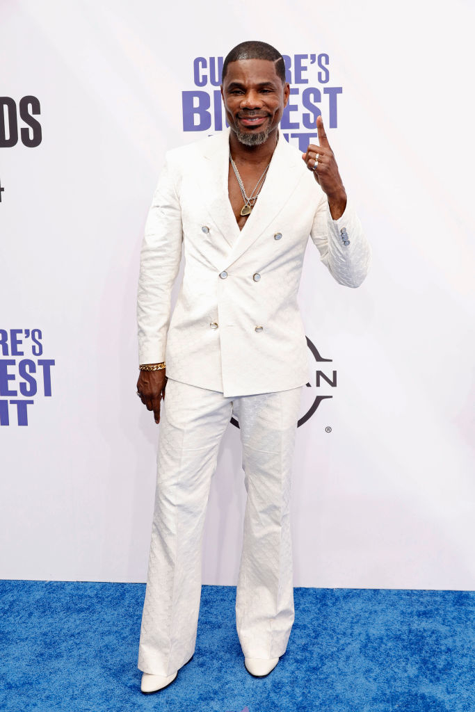 Kirk Franklin on the red carpet wearing a white double-breasted suit, smiling, and pointing upwards with one finger
