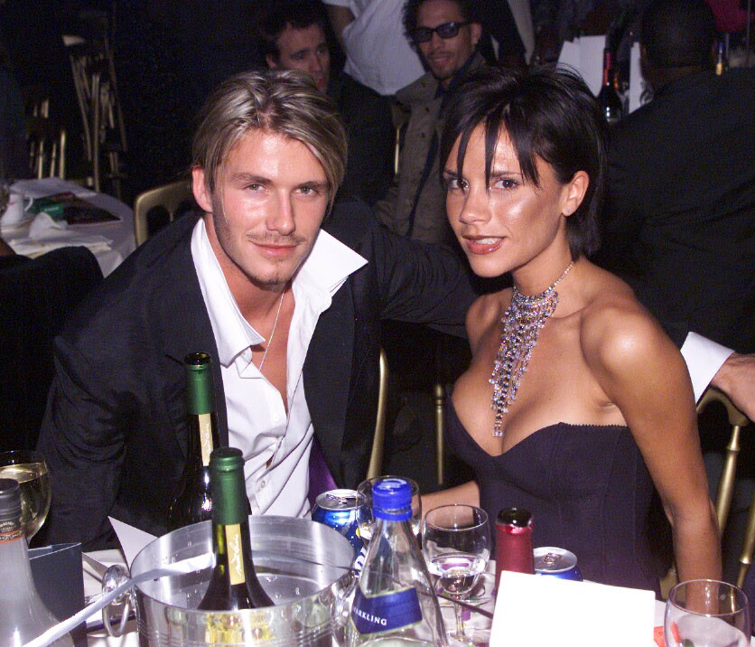 David Beckham and Victoria Beckham sitting at a table, dressed elegantly for an event. David is in a suit, and Victoria is wearing a strapless dress with a statement necklace