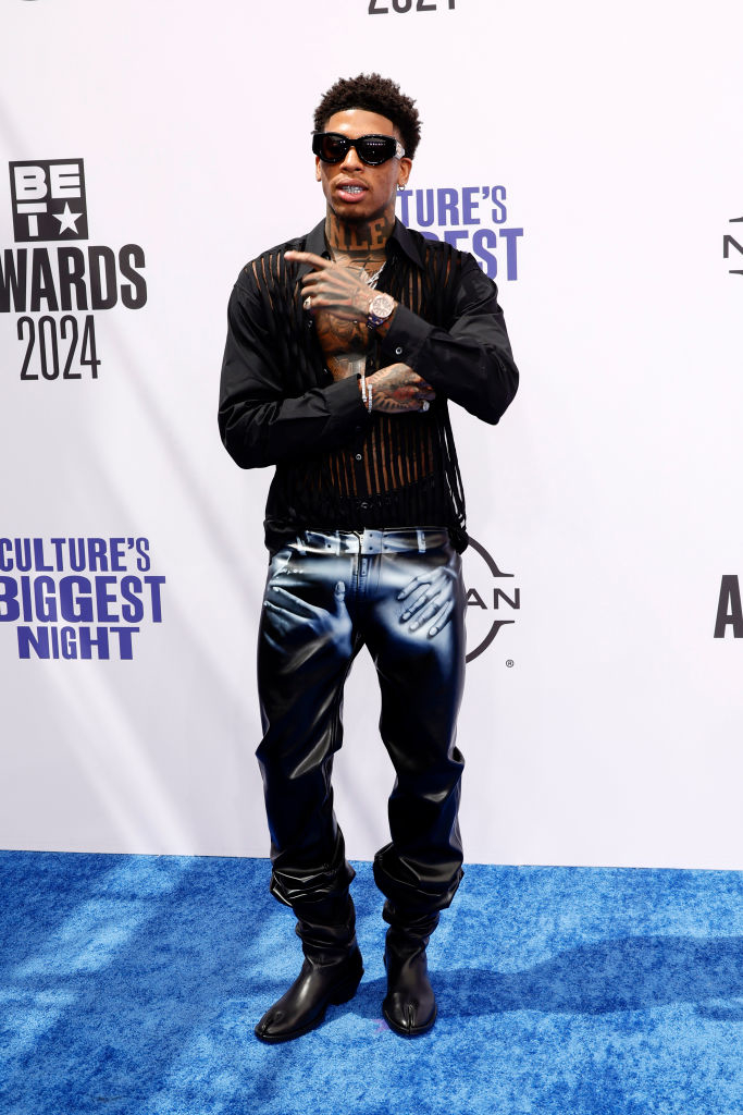 NLE Choppa at the BET Awards 2024, wearing a black mesh shirt with cutouts, leather pants, and sunglasses, on the red carpet