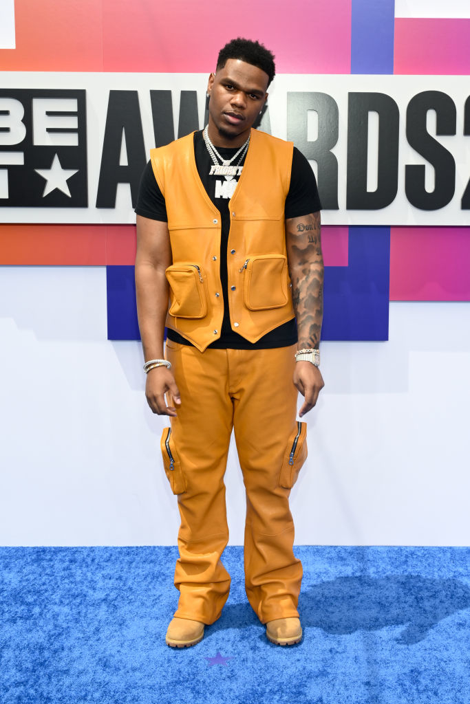 Fridayy at the BET Awards in a black t-shirt, orange leather vest with matching pants, and tan boots. He wears multiple silver chain necklaces