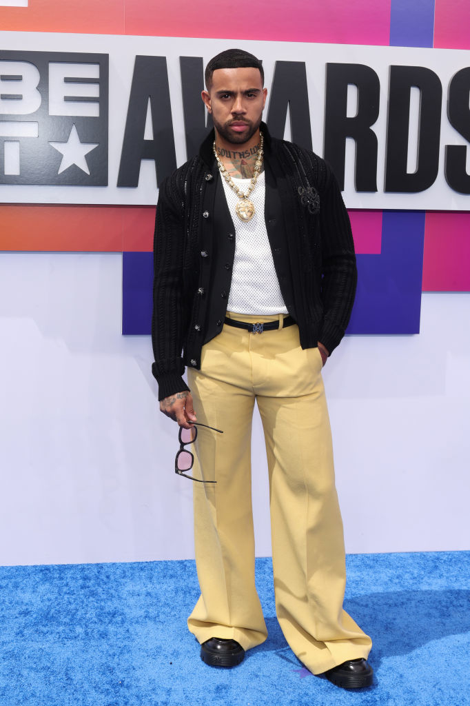Vic Mensa on the red carpet at the BET Awards, wearing a white shirt, black sweater, and yellow pants, holding eyeglasses