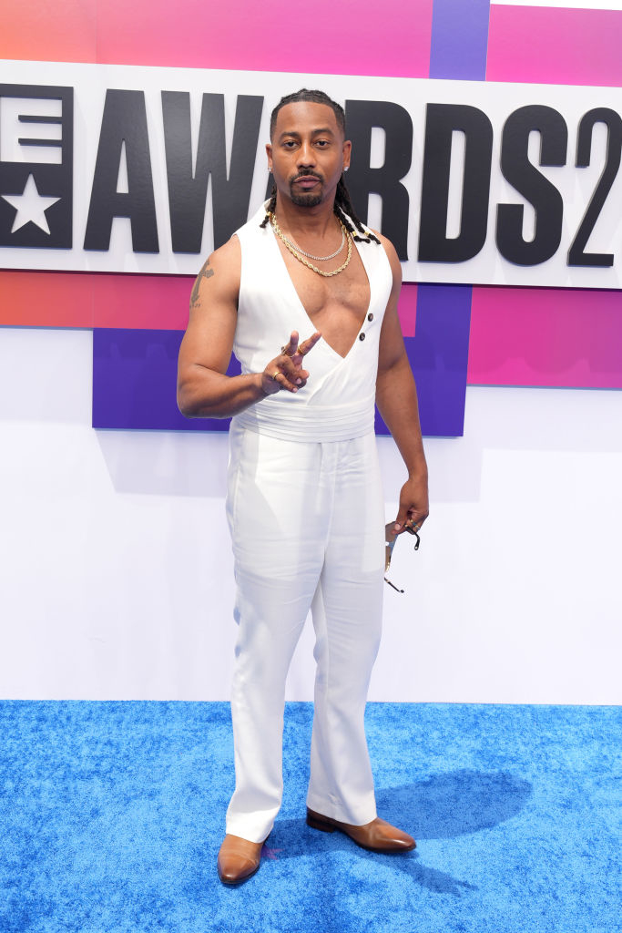Brandon T. Jackson poses on a blue carpet at an awards event, wearing a white sleeveless jumpsuit with a deep V-neck, holding sunglasses and making a peace sign