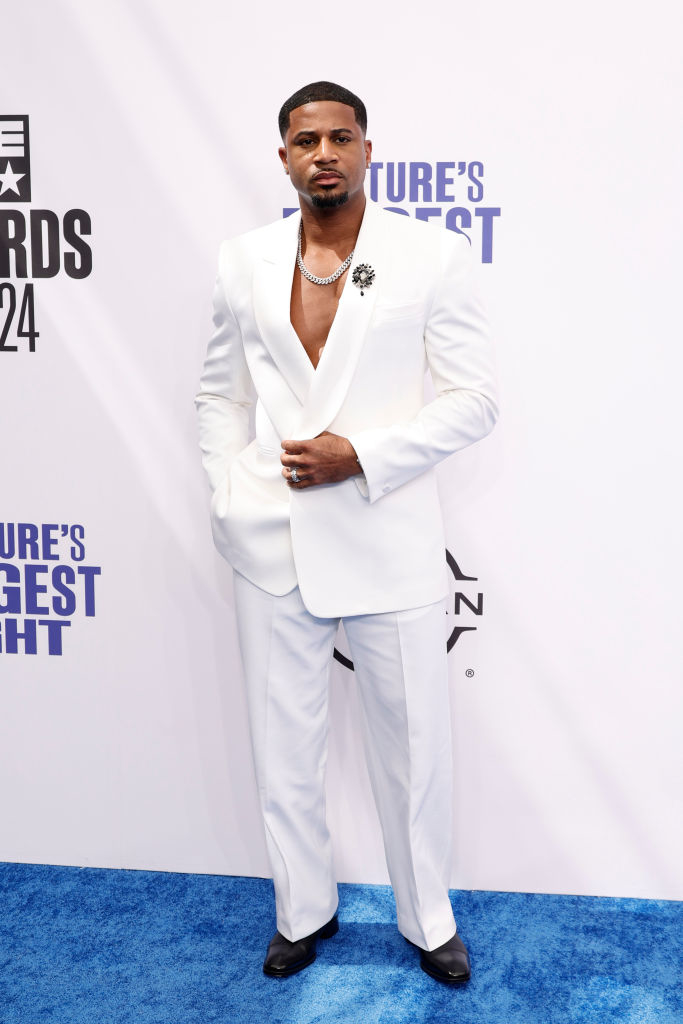 Devale Ellis in a white suit with a deep V-neck shirt on the blue carpet at an awards show