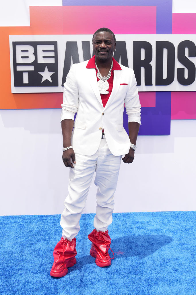 Akon on the red carpet at the BET Awards, wearing a white suit with red accents and large red boots, accessorized with a large silver necklace
