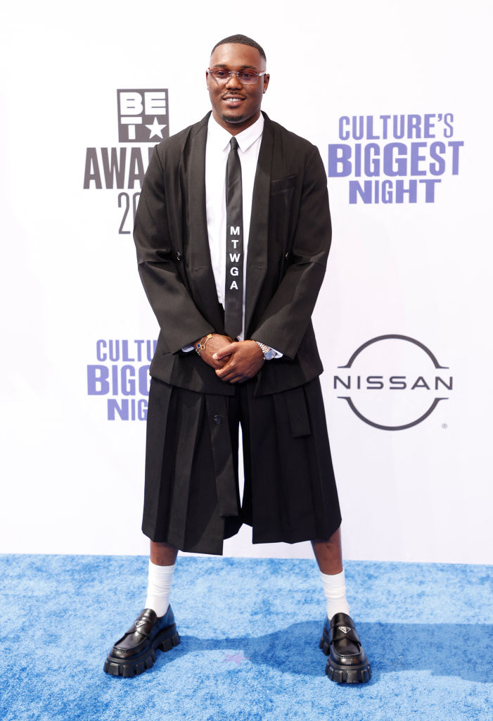 Kalan.FrFr stands on the blue carpet at the BET Awards 2021 wearing a black suit with shorts, a tie labeled "MTWGA," and chunky black shoes