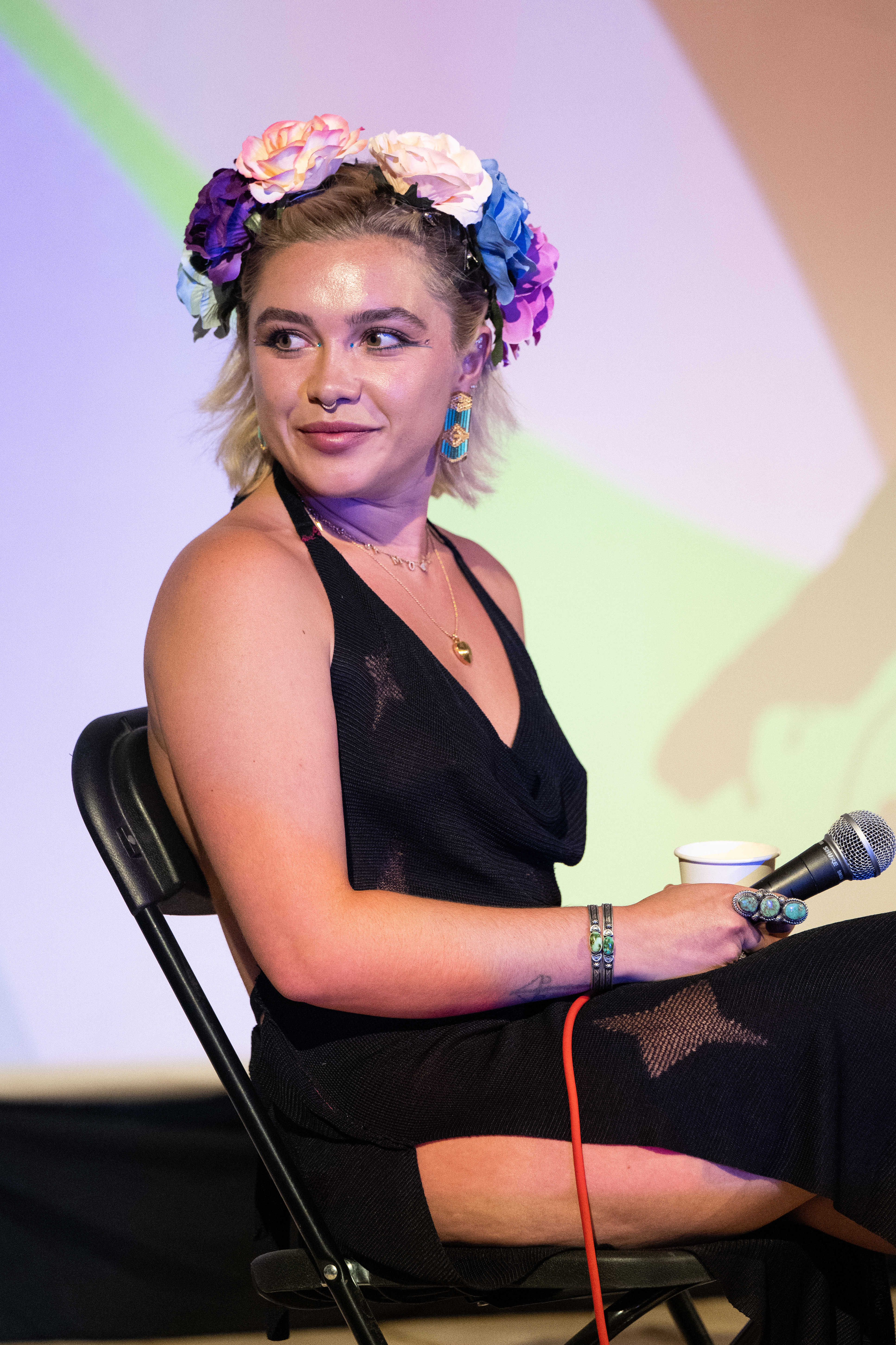 Florence Pugh sits on a chair, wearing a black dress with a deep neckline and slits, a colorful flower crown, and holding a microphone and cup