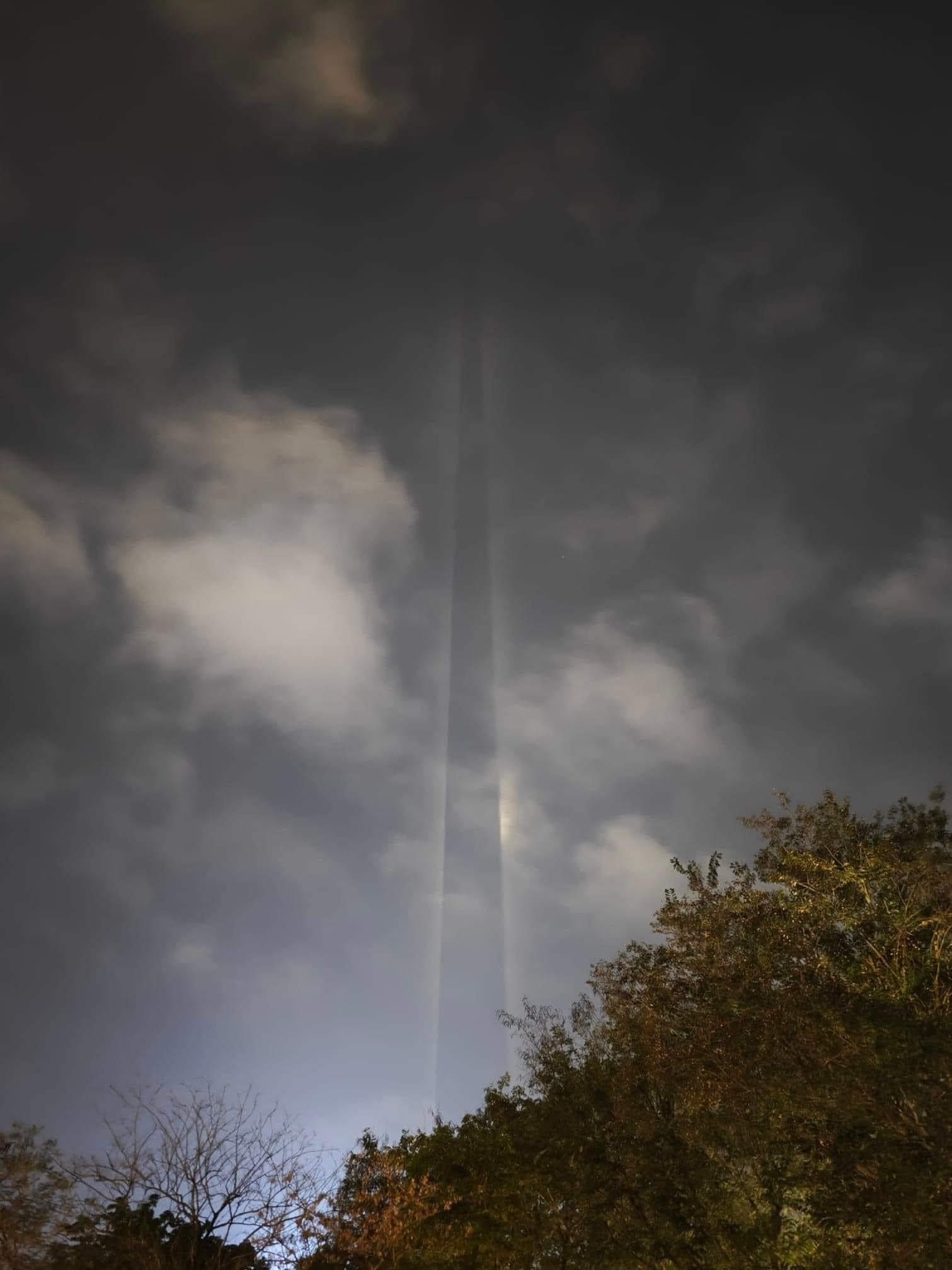 A tall, thin vertical beam of light extends into the cloudy night sky