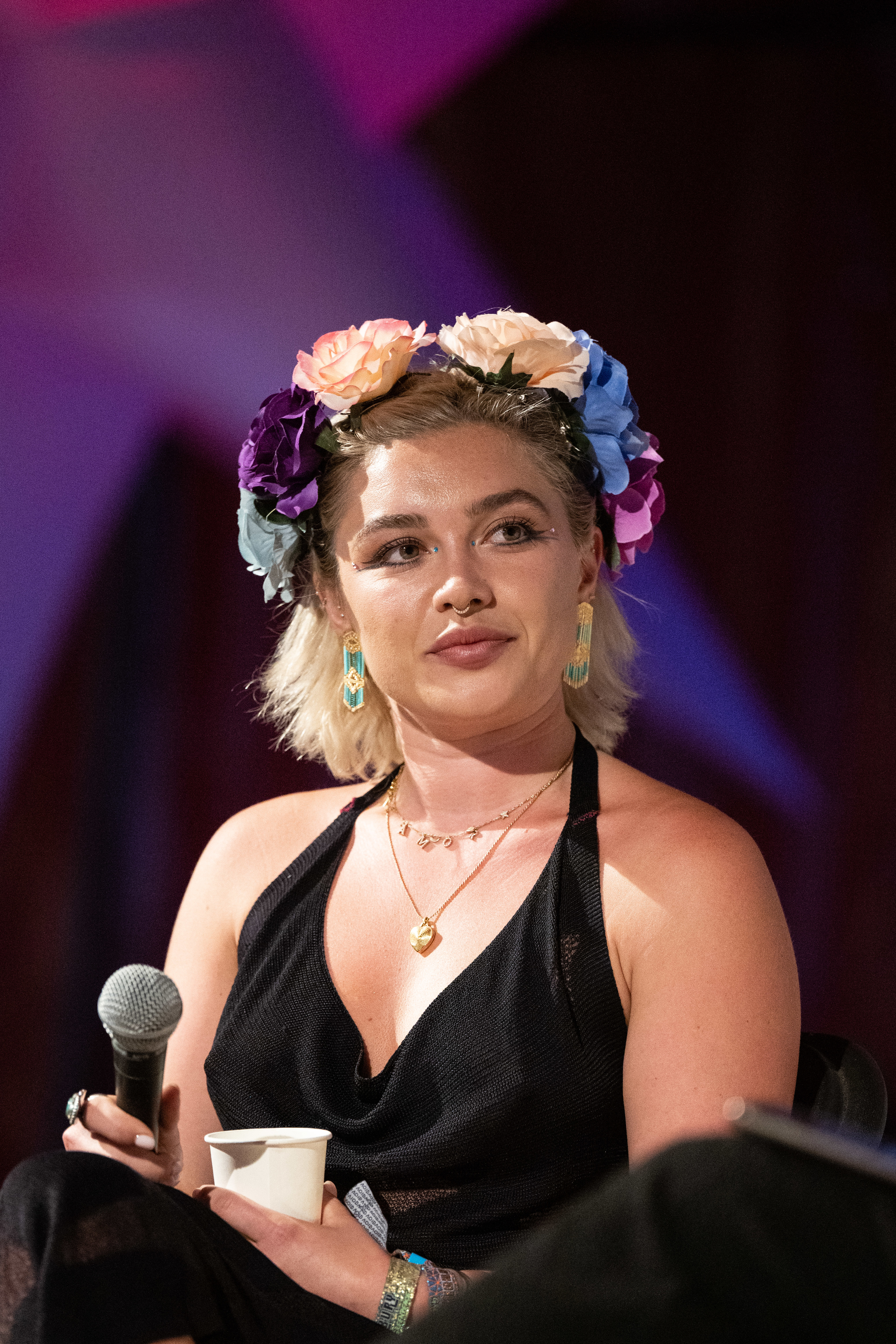 Florence Pugh is sitting and holding a microphone, wearing a floral headband, black halter top, and gold jewelry. She has a calm expression on her face