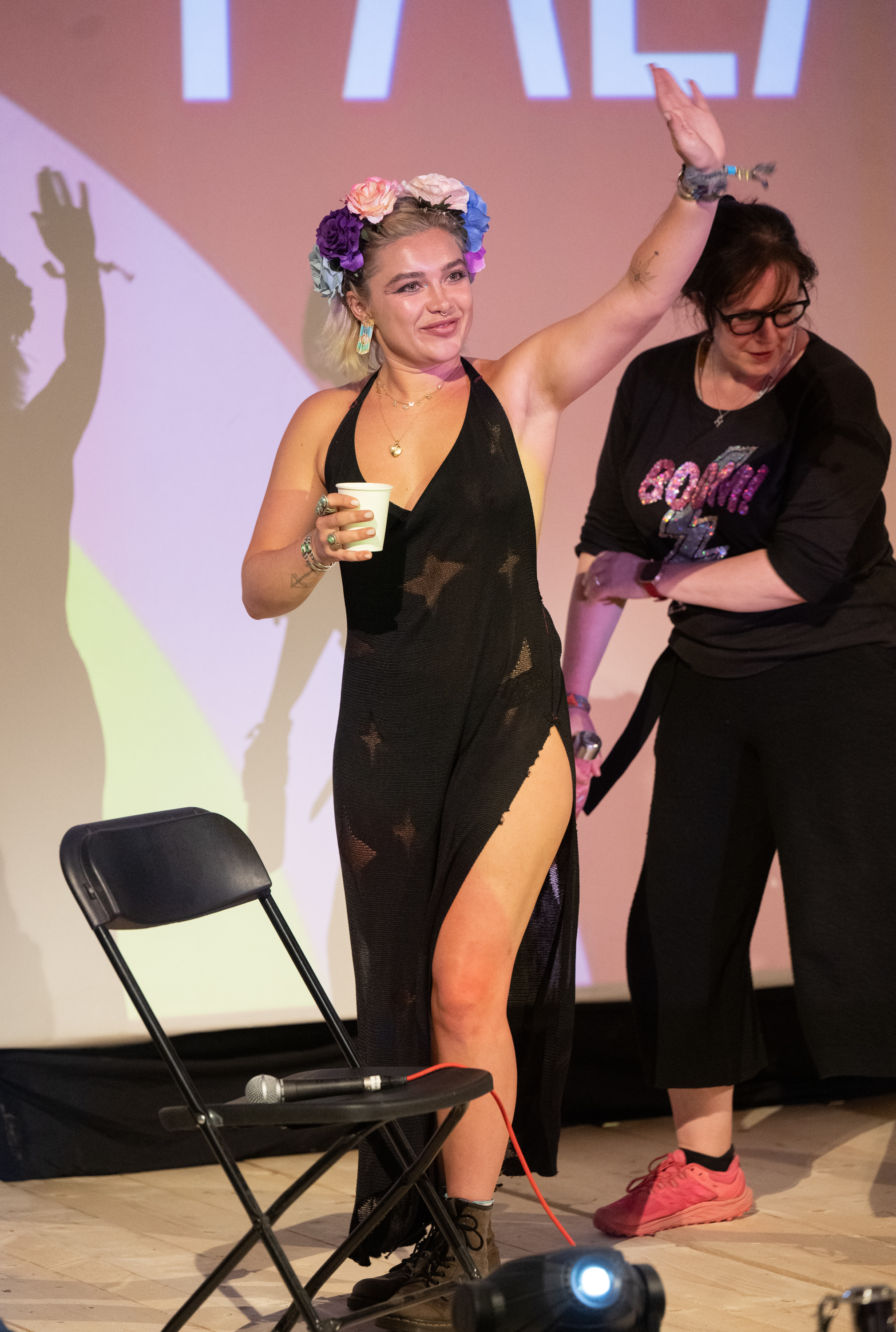 Florence Pugh waves on stage wearing a floral crown and a sleeveless black dress. A seated microphone and an unidentified woman are also in the image