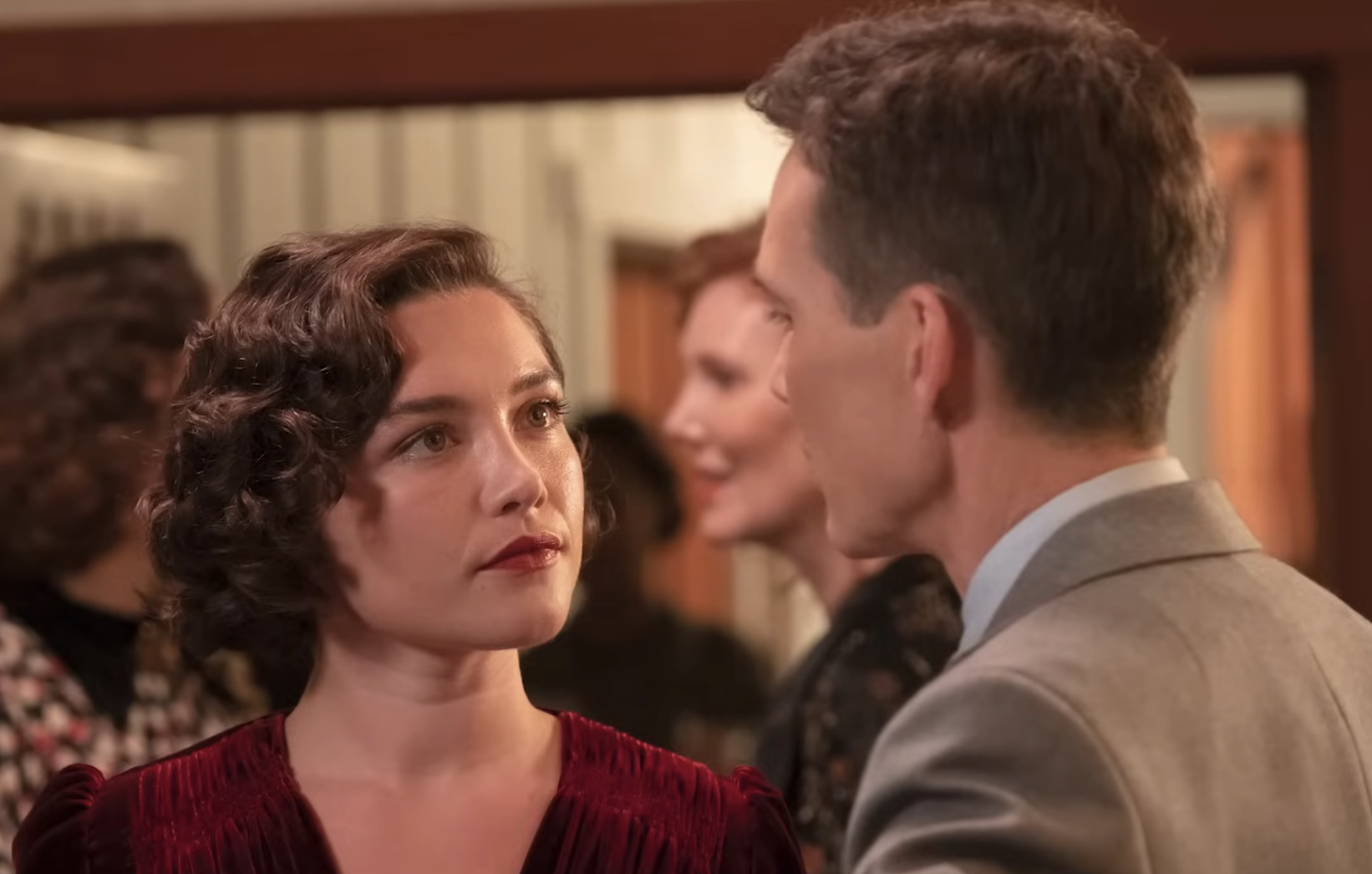 Florence Pugh and a man in a suit having a serious conversation at a social event, with two women chatting in the background