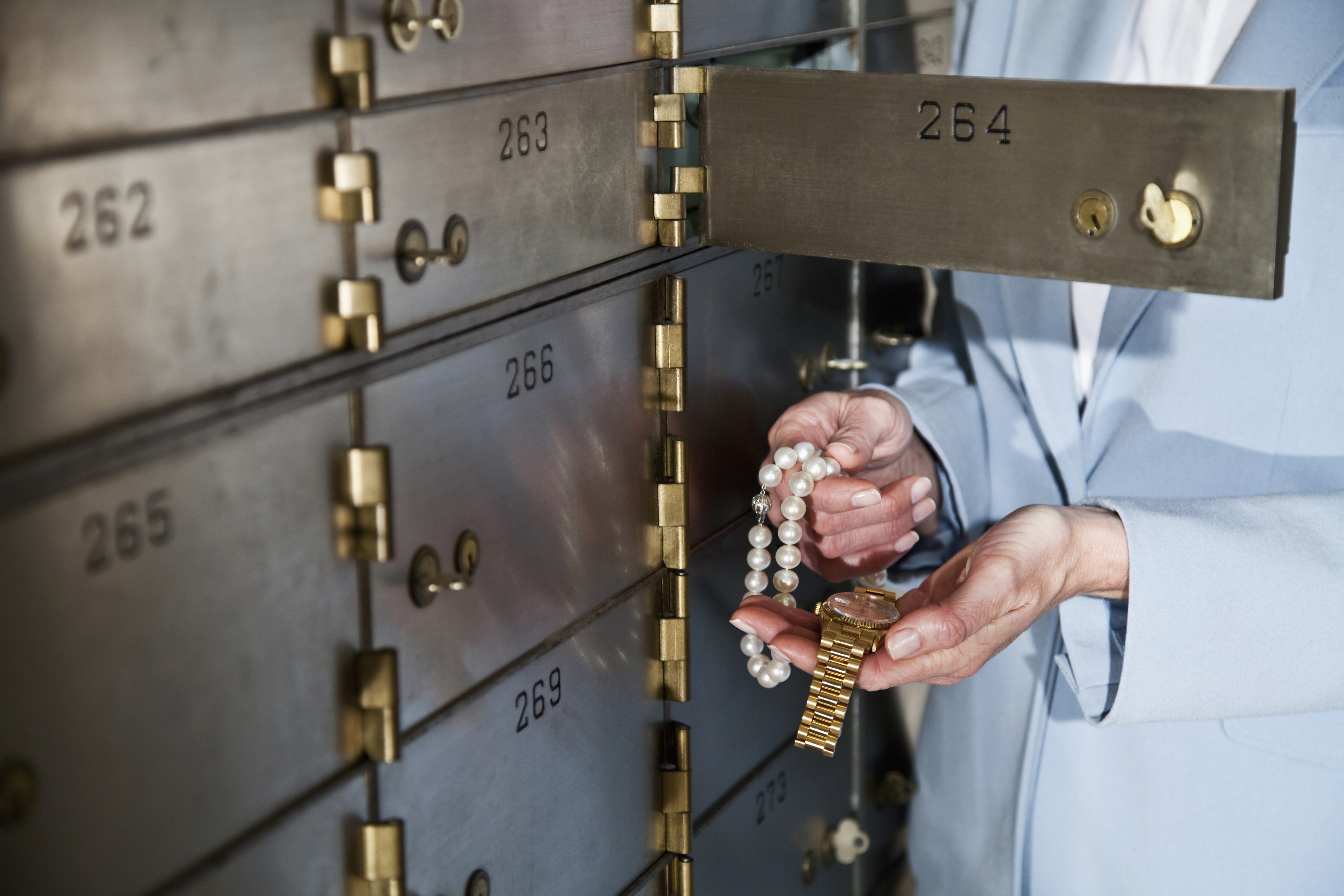 A person in a suit holds a gold watch and a pearl necklace, standing in front of open safety deposit boxes in a vault