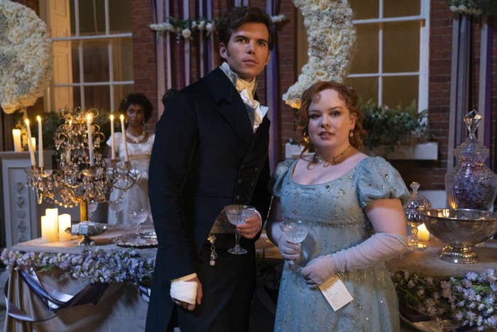 Luke Newton and Nicola Coughlan dressed in Regency-era attire, holding champagne glasses at an elegant indoor gathering with candles and floral decorations in a scene from Bridgerton