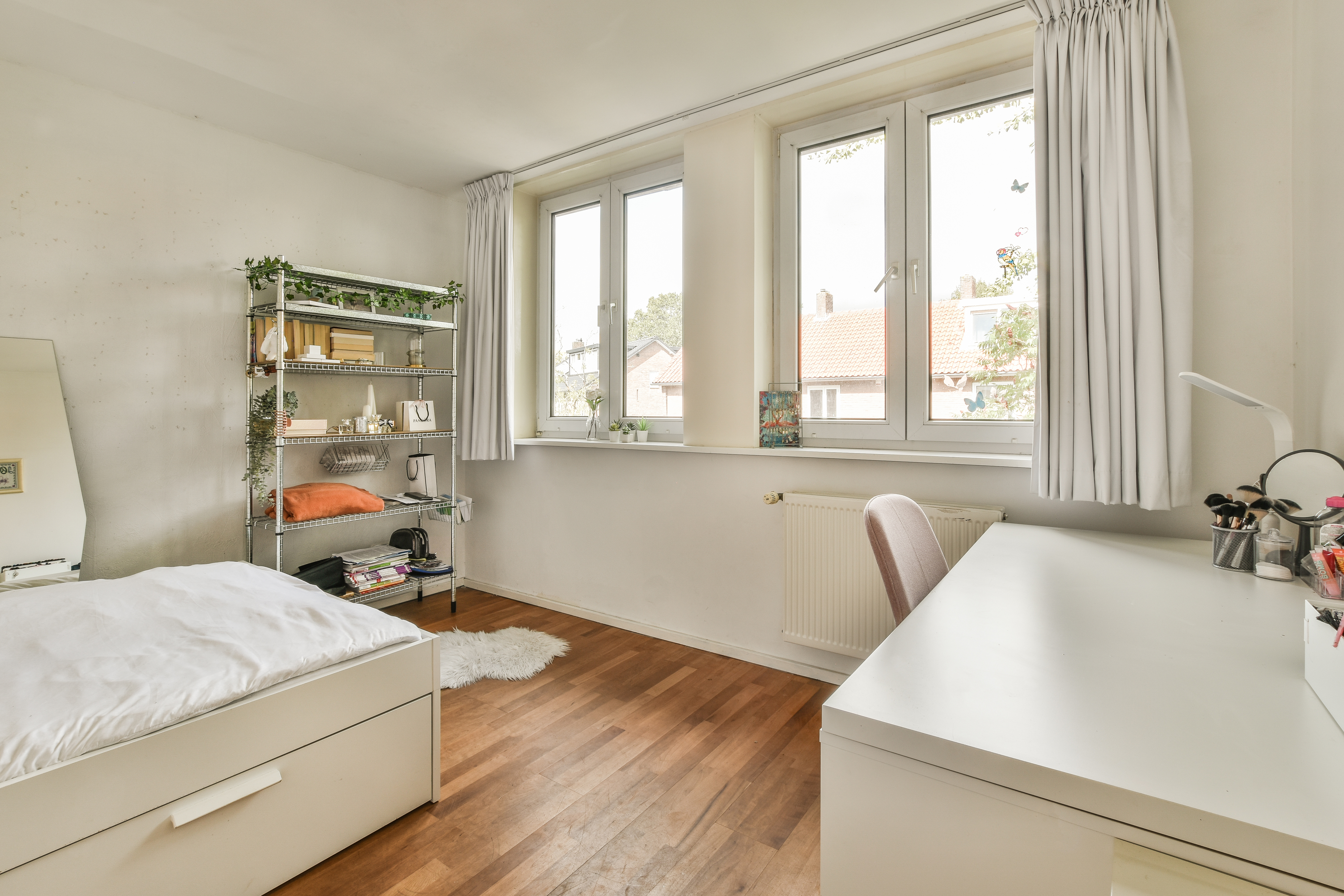 A bright, neatly organized home office/bedroom with a desk, chair, bed, and a metal shelving unit against the wall, featuring a large window allowing ample natural light