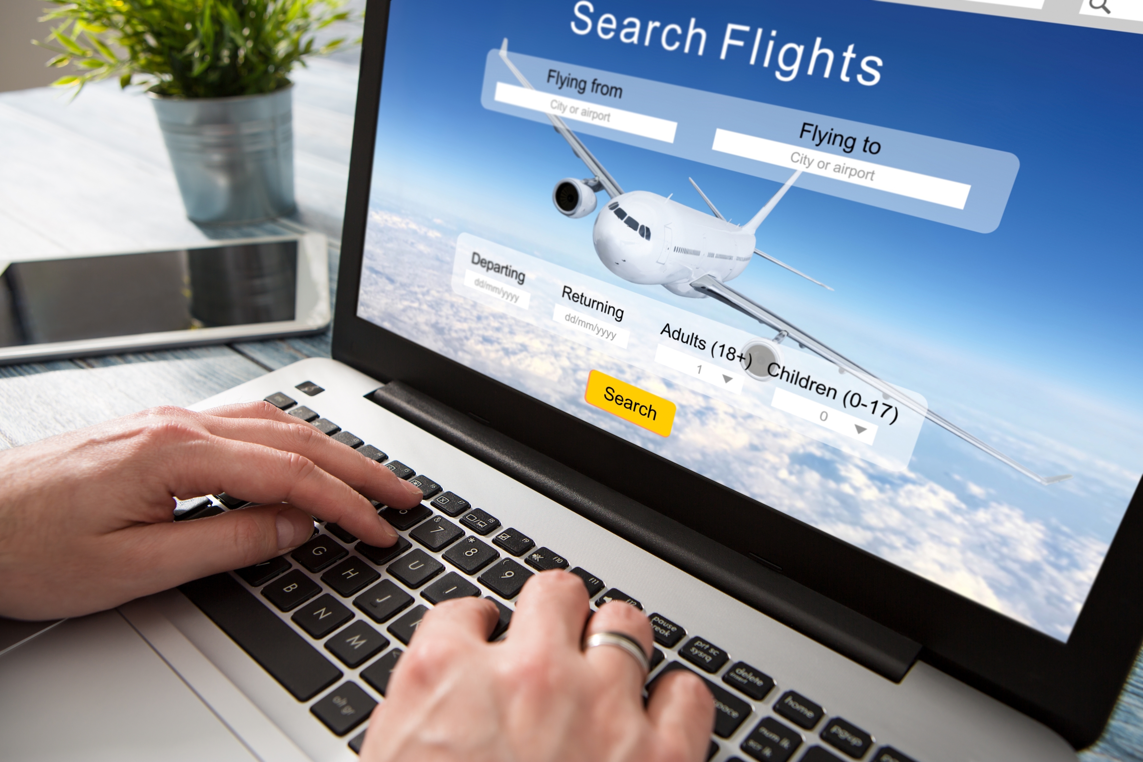 A person using a laptop with a flight search website on the screen. The search fields include flying from, flying to, departing, returning, adults, and children