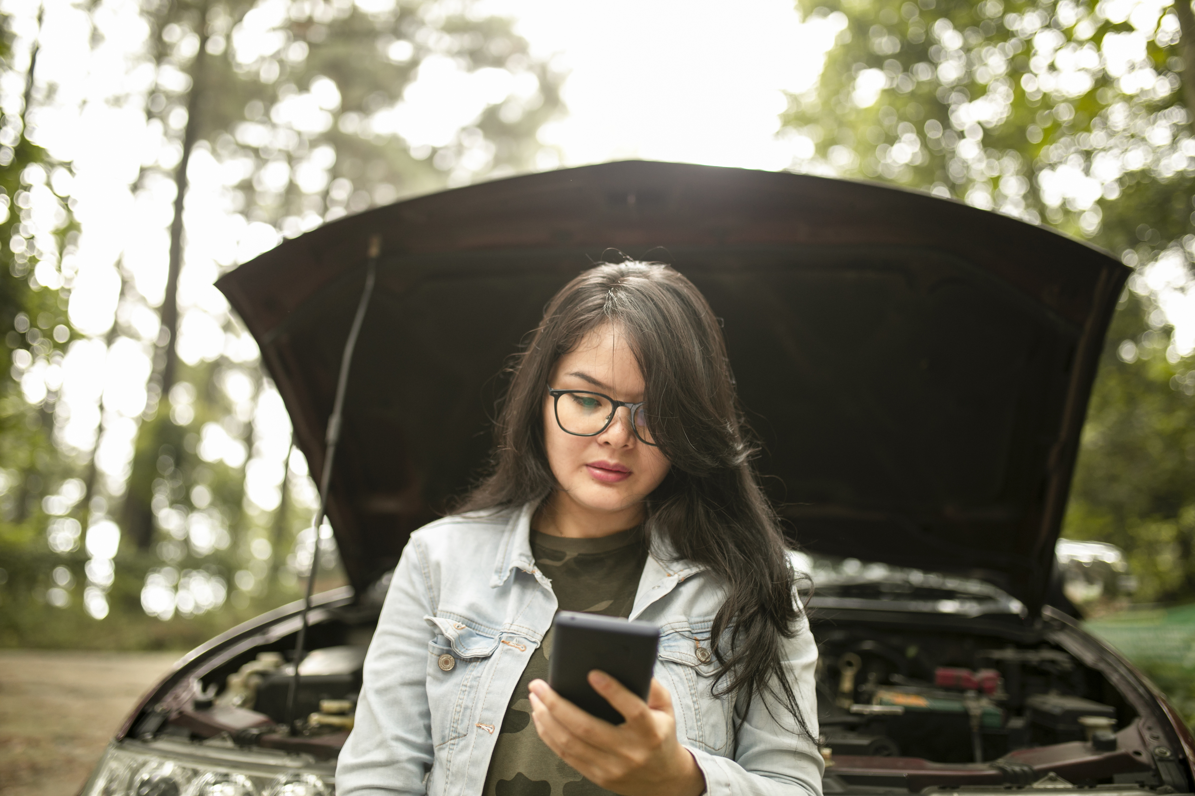A person stands in front of a car with the hood up, looking at their phone. The image relates to work and money and suggests a car breakdown situation